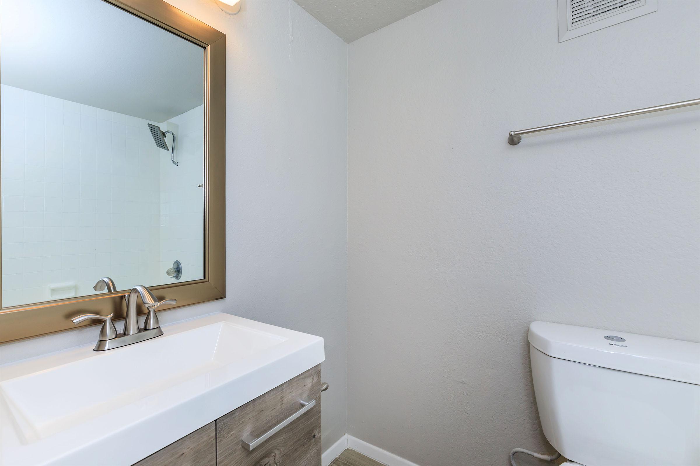 A modern bathroom featuring a large mirror above a sink with two faucets and wooden cabinets. To the right, there is a white toilet. The walls are painted light gray, and there's a shower area visible in the background. The overall design is clean and minimalist.