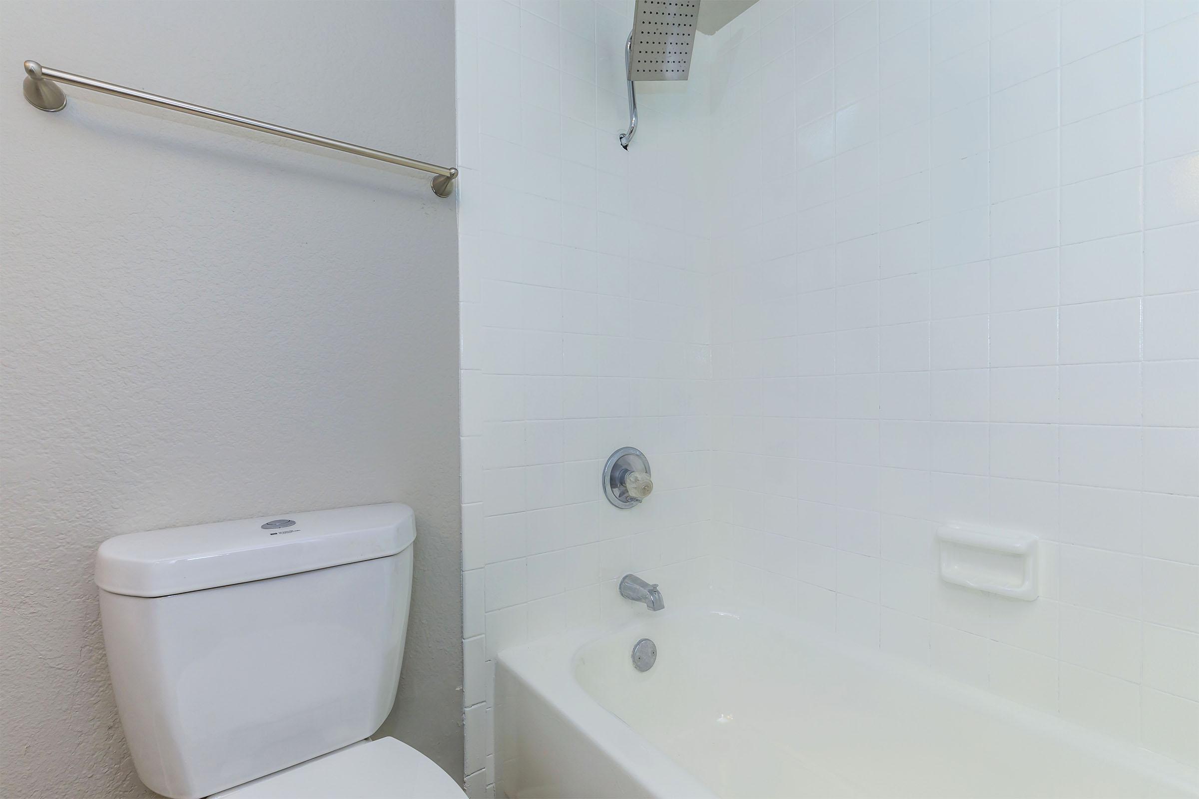 A clean bathroom featuring a white tiled shower and bathtub. The toilet is visible to the left, with a simple metal grab bar mounted on the wall. The overall aesthetic is minimalistic and well-maintained.