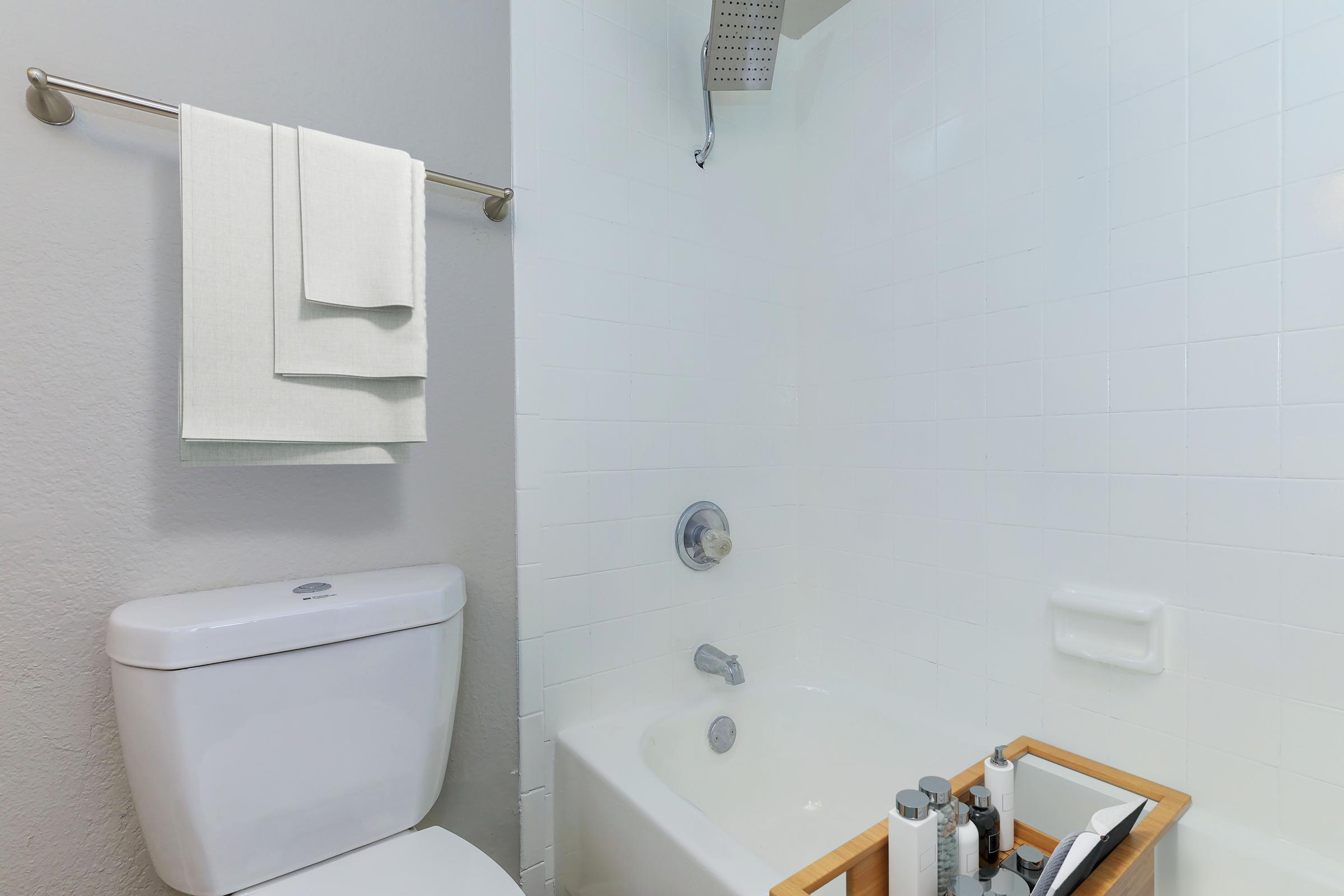 A clean and modern bathroom featuring a white bathtub, a wall-mounted showerhead, and a toilet. Two neatly hung towels are on a towel bar. A wooden caddy holds various toiletries on the tub's edge, and the walls are painted a light gray with shiny white tiles.