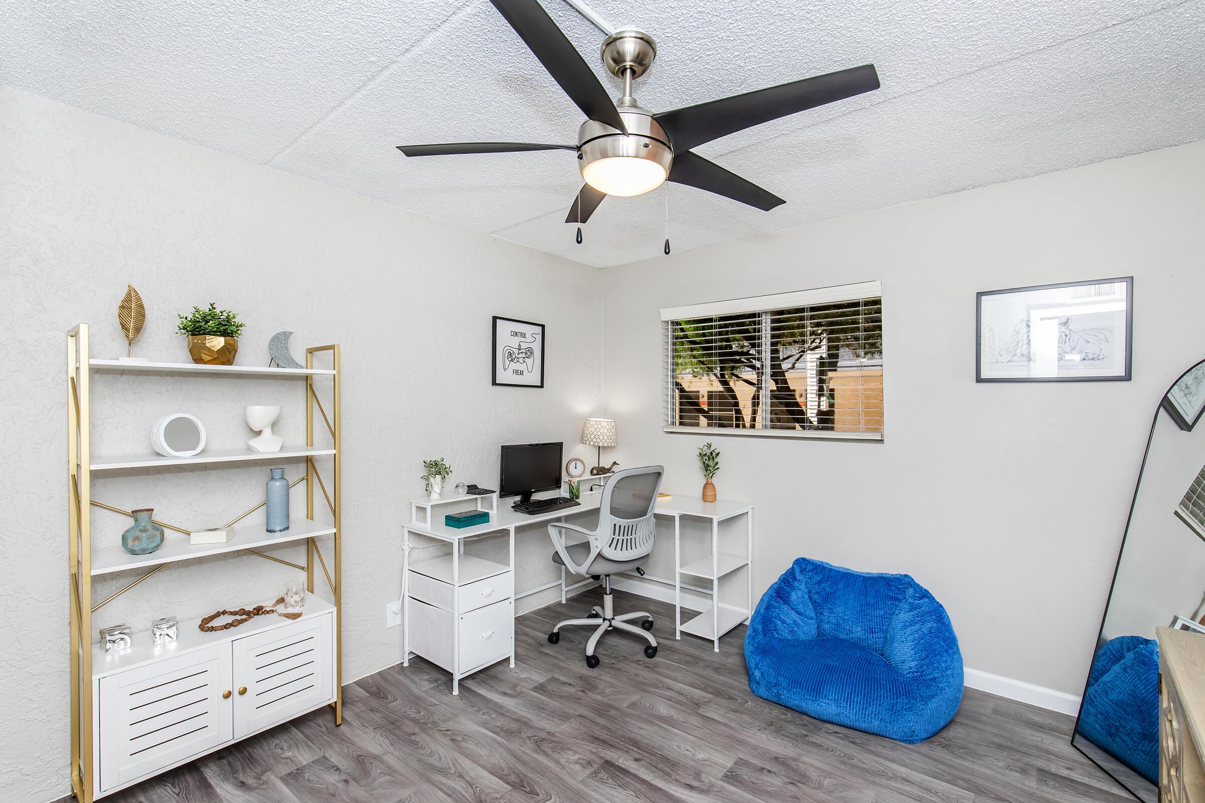 A modern home office featuring a white desk with a computer, a grey chair, and a bookshelf with decorative items. There's a cozy blue bean bag in the corner and a large mirror on the right. A ceiling fan is overhead, and the room has natural light from a window. The walls are light grey with wooden flooring.