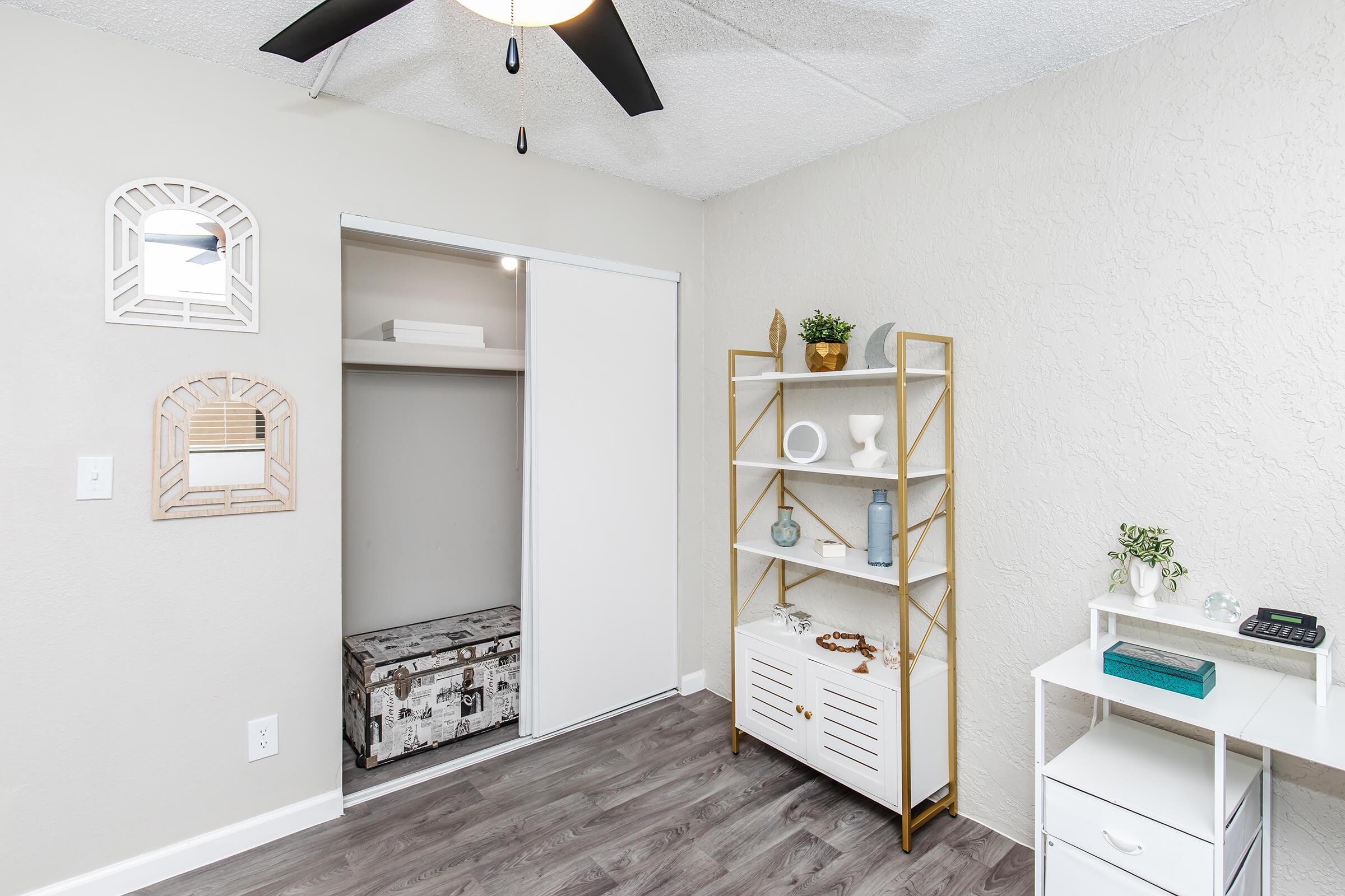 A well-lit bedroom featuring a ceiling fan, a closed sliding closet door, a decorative bookshelf with vases and plants, and a stylish white cabinet. The walls are painted light gray, and the flooring is wooden with a modern aesthetic.