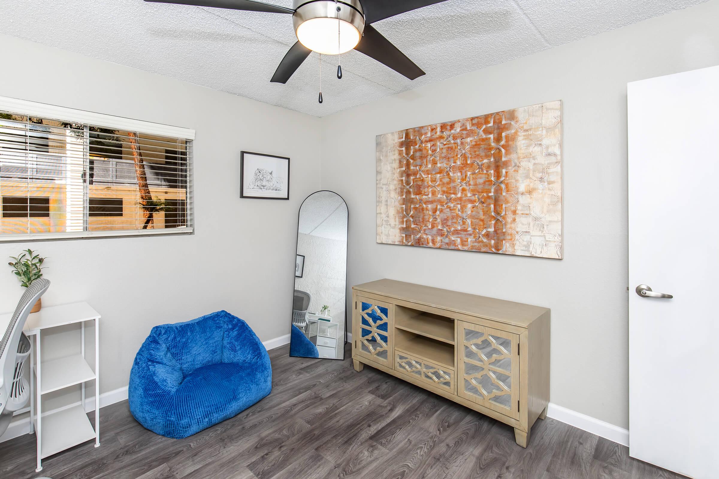A bright, modern bedroom featuring a blue bean bag, a large mirror, and a decorative wooden cabinet. The walls are light-colored with a textured artwork hanging above the cabinet. Natural light streams in through a window, and a ceiling fan is visible above. The flooring is a dark laminate.