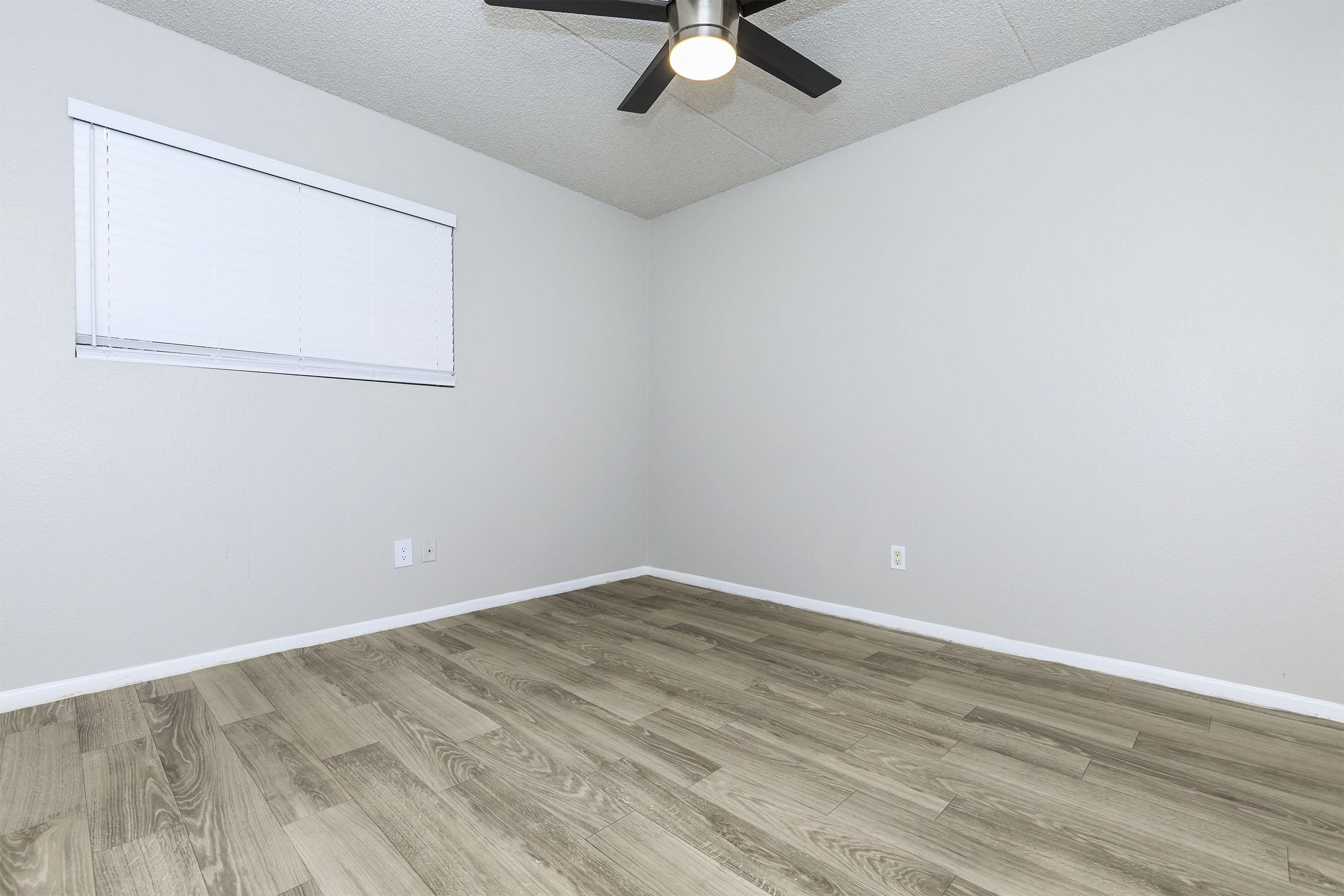 Empty room with light gray walls and a ceiling fan. A window with white blinds allows natural light in, and the floor is covered with light wood laminate. The space is uncluttered, providing a blank canvas for potential furniture arrangement.