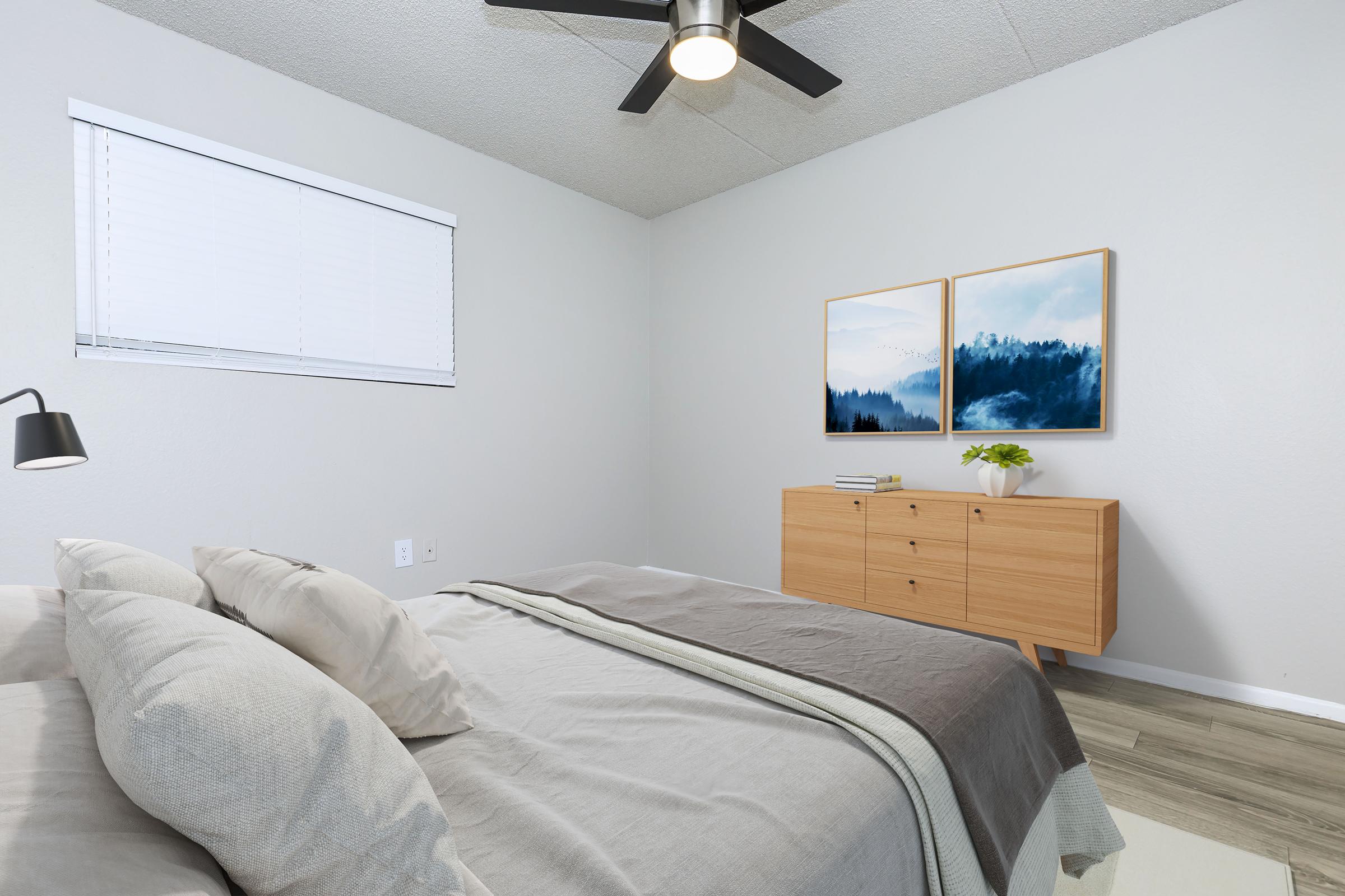 A cozy, modern bedroom featuring a neatly made bed with gray linens, a wooden dresser, and two framed landscape paintings on the wall. Natural light streams in through a window with white blinds, and a ceiling fan adds a contemporary touch to the serene atmosphere.