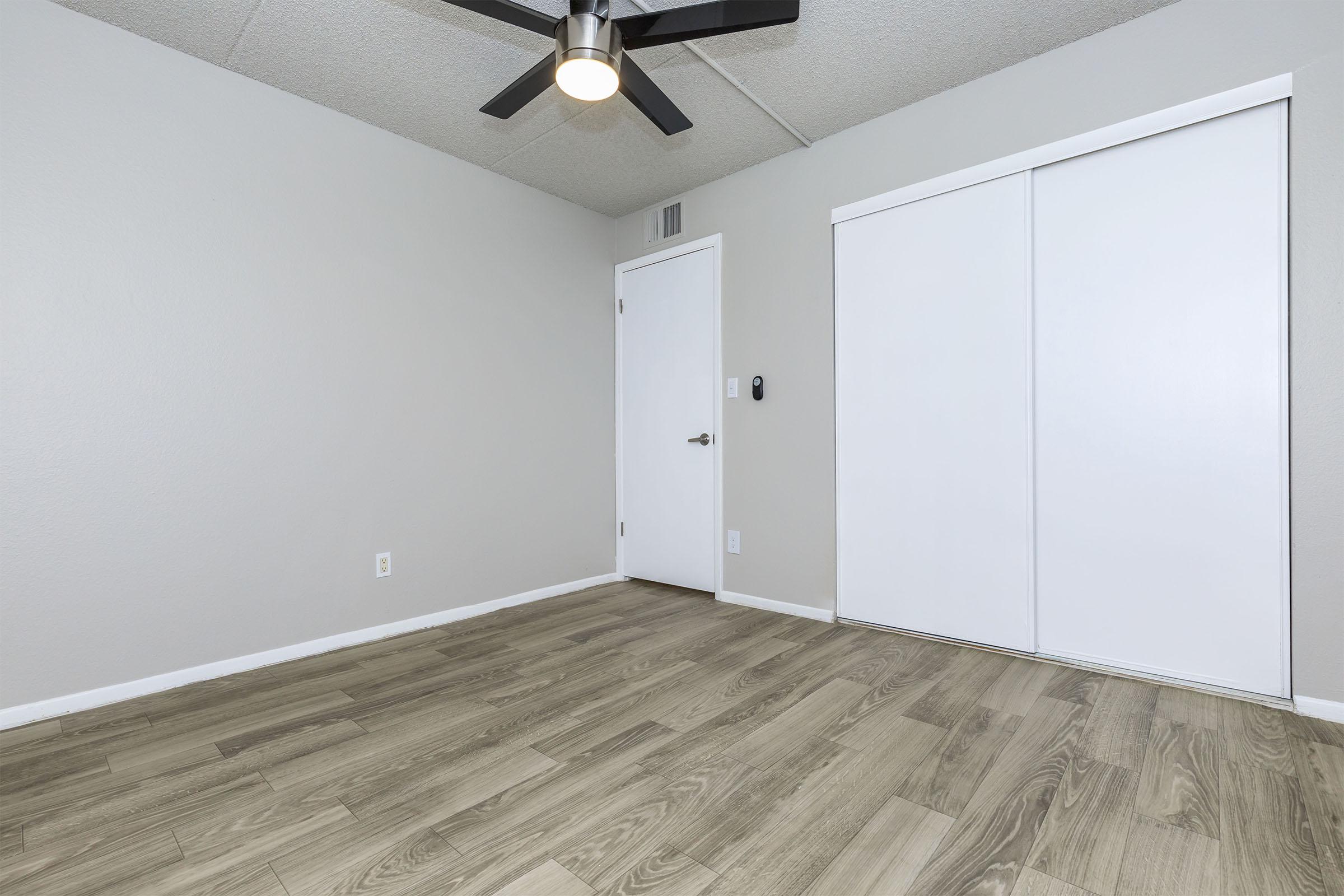 A minimalist interior room with light gray walls and a wooden floor. There is a white sliding closet door on the right and a closed door on the left leading to another space. A ceiling fan with a light fixture is mounted in the center of the room, which has an overall clean and modern aesthetic.