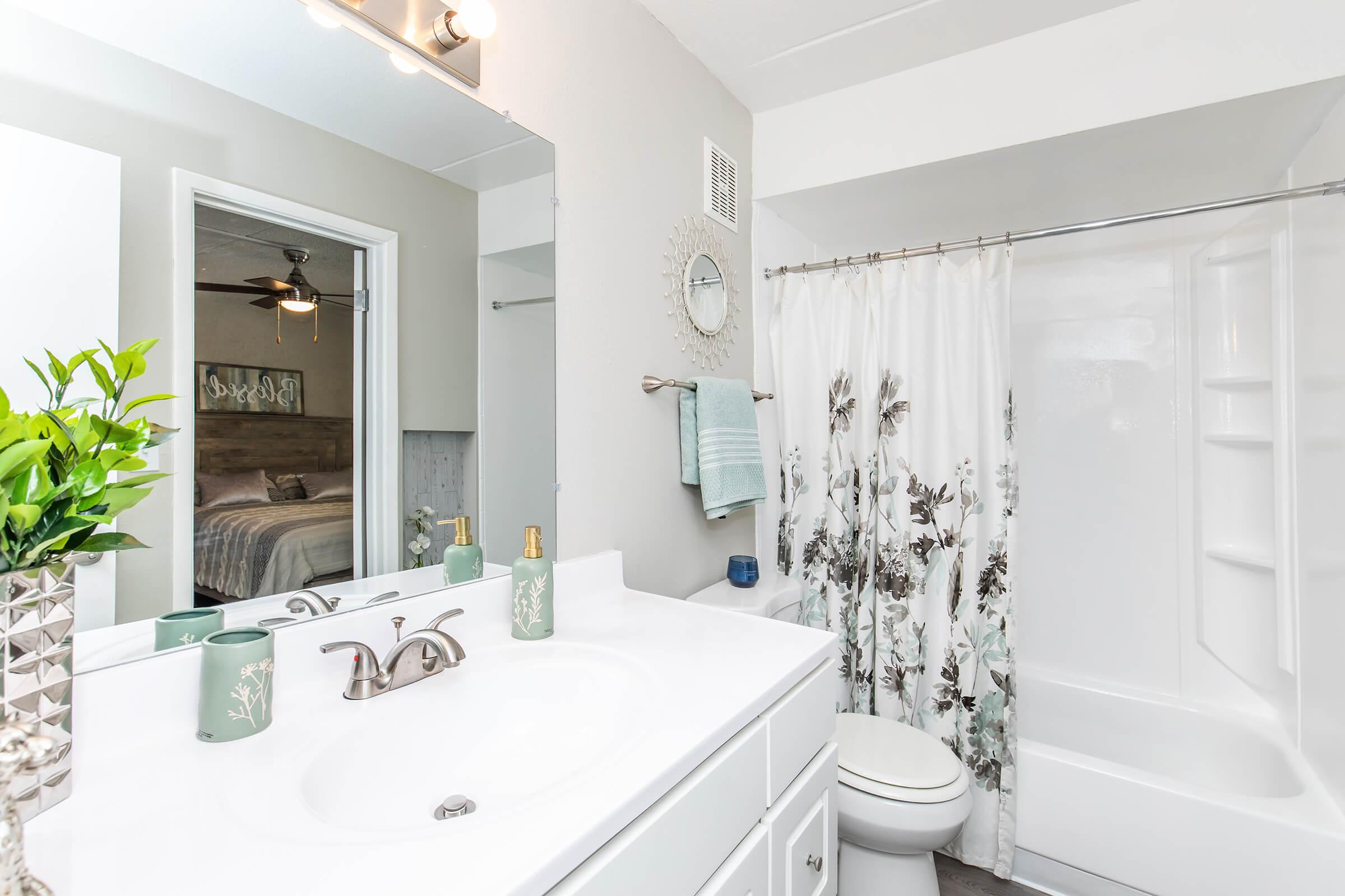 A modern bathroom featuring a white vanity with a sink, a mirror above it, and a shower/tub combination. The shower curtain has a floral design, and there are decorative items like a potted plant and toiletries on the counter. A light fixture is mounted above the mirror, and the wall is painted a light neutral color.