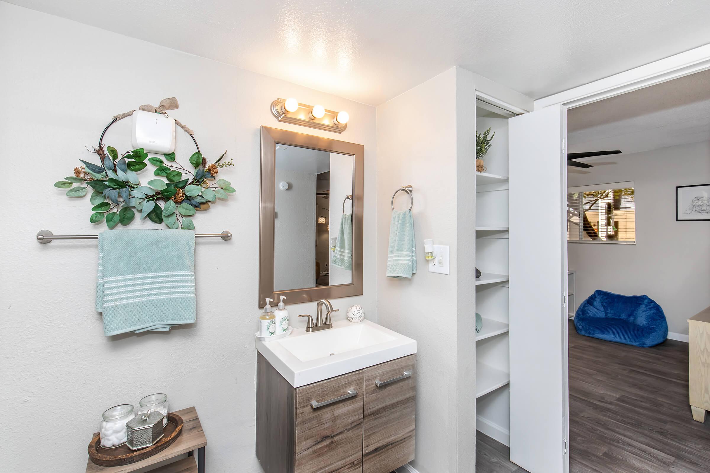 A modern bathroom featuring a light wood vanity with a white sink, a large mirror framed in wood, and a decorative wall wreath with greenery. There are aqua-colored towels hanging on a rack, and a few decorative items on the counter. A doorway leads to another room with a blue bean bag chair and a light wood table.