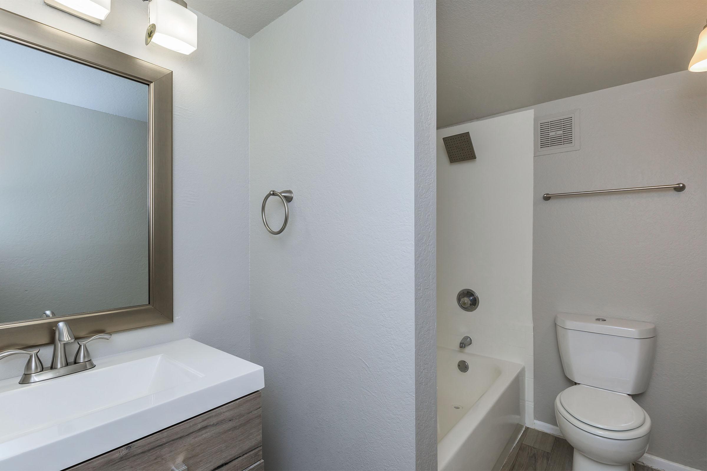 Modern bathroom featuring a rectangular mirror above a white sink with a sleek cabinet below. A bathtub is visible on the left, with a showerhead above. The walls are painted light gray, and a towel ring is mounted beside the sink. A toilet is located on the right side, with a simple light fixture above.