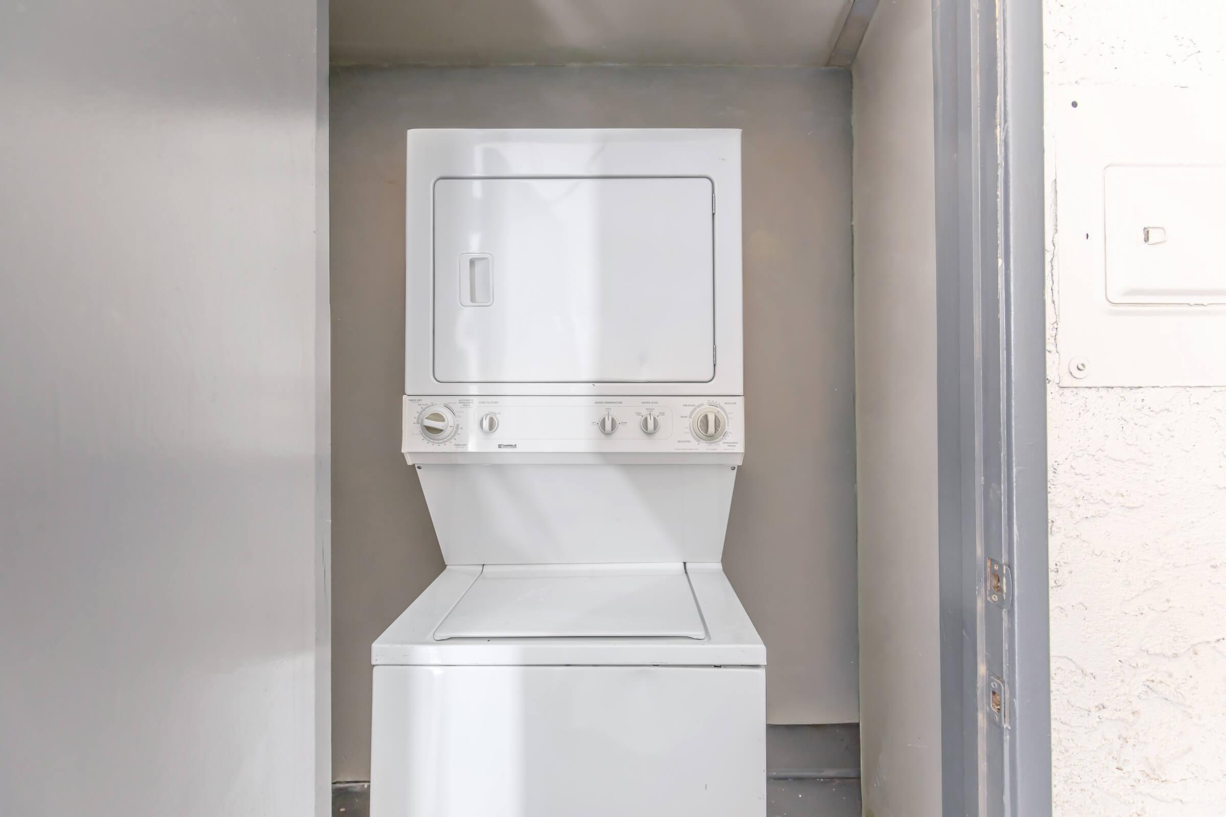 A stacked washer and dryer unit in a small laundry nook. The walls are painted a neutral color, and there is a power outlet on the right side. The appliances have a white finish, and the room appears clean and organized.