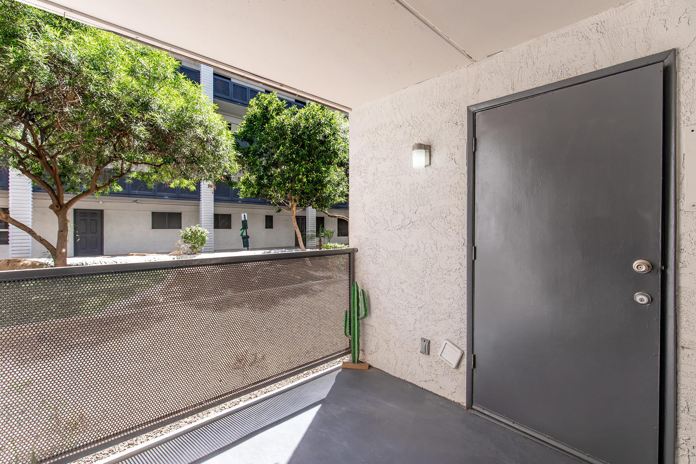 A view of a modern apartment entrance featuring a gray door, a metal railing, and a small cactus decoration. In the background, green trees and adjacent buildings are visible, creating a pleasant outdoor setting. The area is well-lit and captures a homey atmosphere.