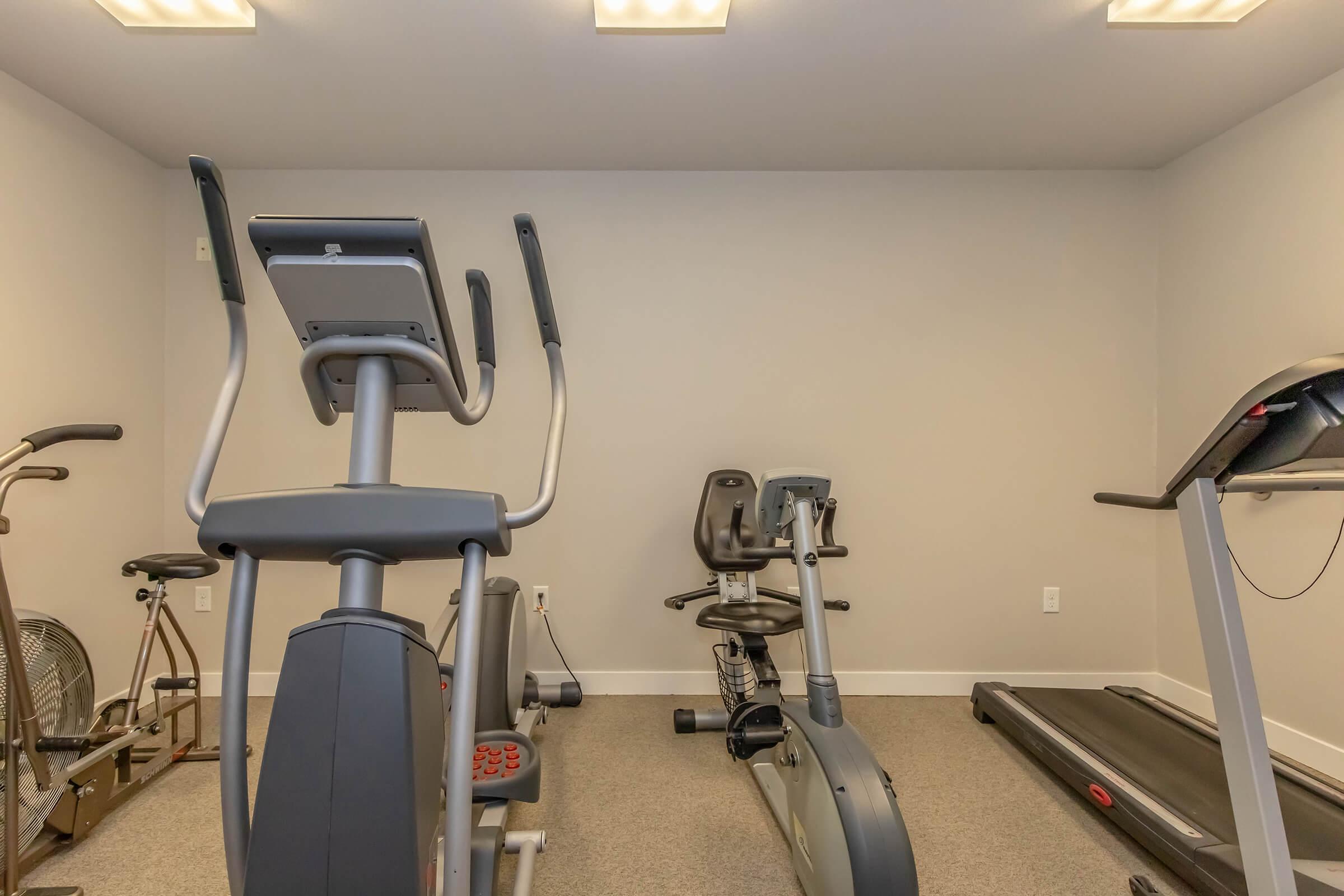 A well-lit home gym featuring various exercise equipment, including an elliptical machine, a stationary bike, and a treadmill, set against a neutral wall color and carpeted flooring.