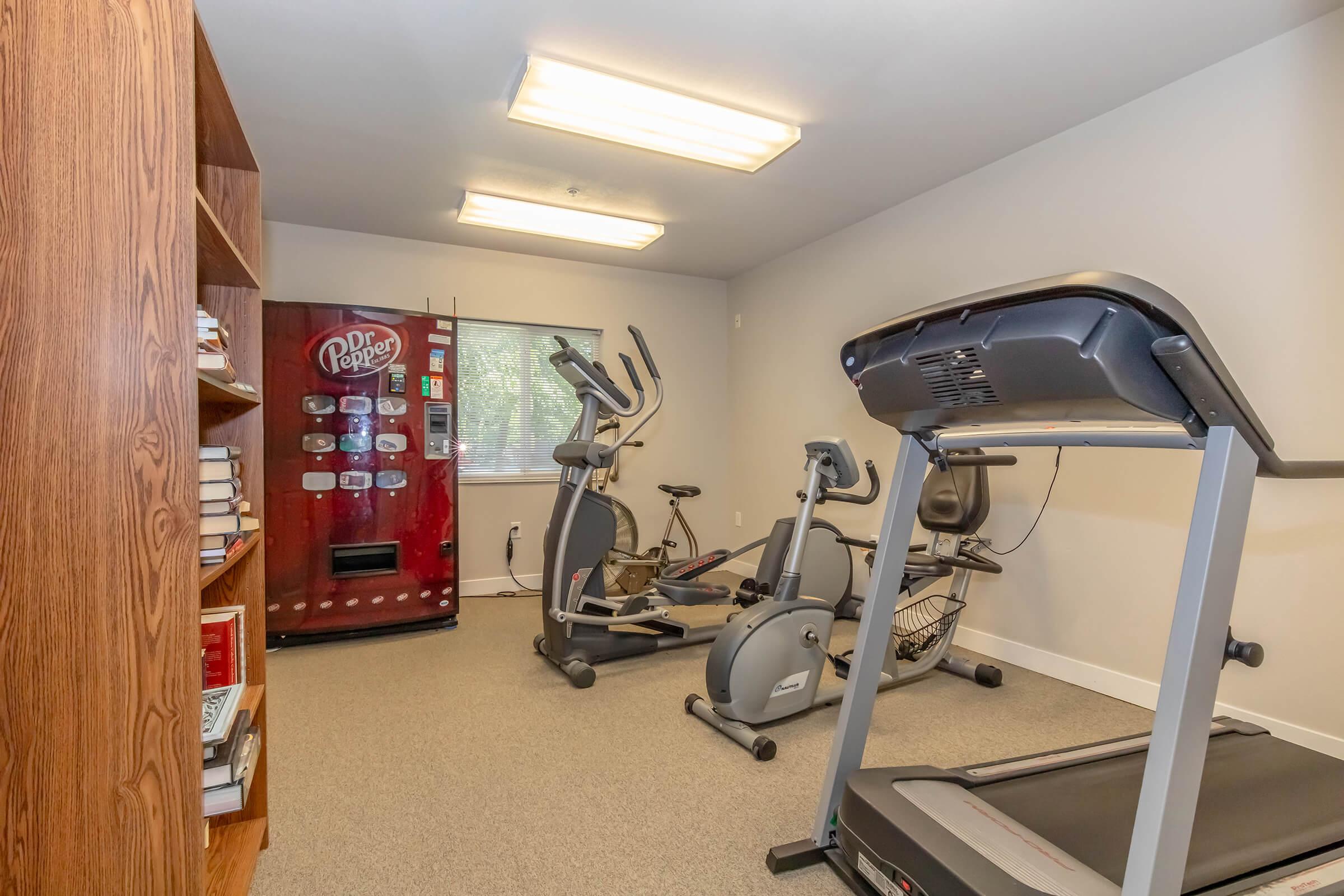 A workout room featuring exercise equipment, including an elliptical machine and treadmill, alongside a bookshelf filled with books. A Dr Pepper vending machine is visible against the wall, and the room is well-lit with overhead fluorescent lights.