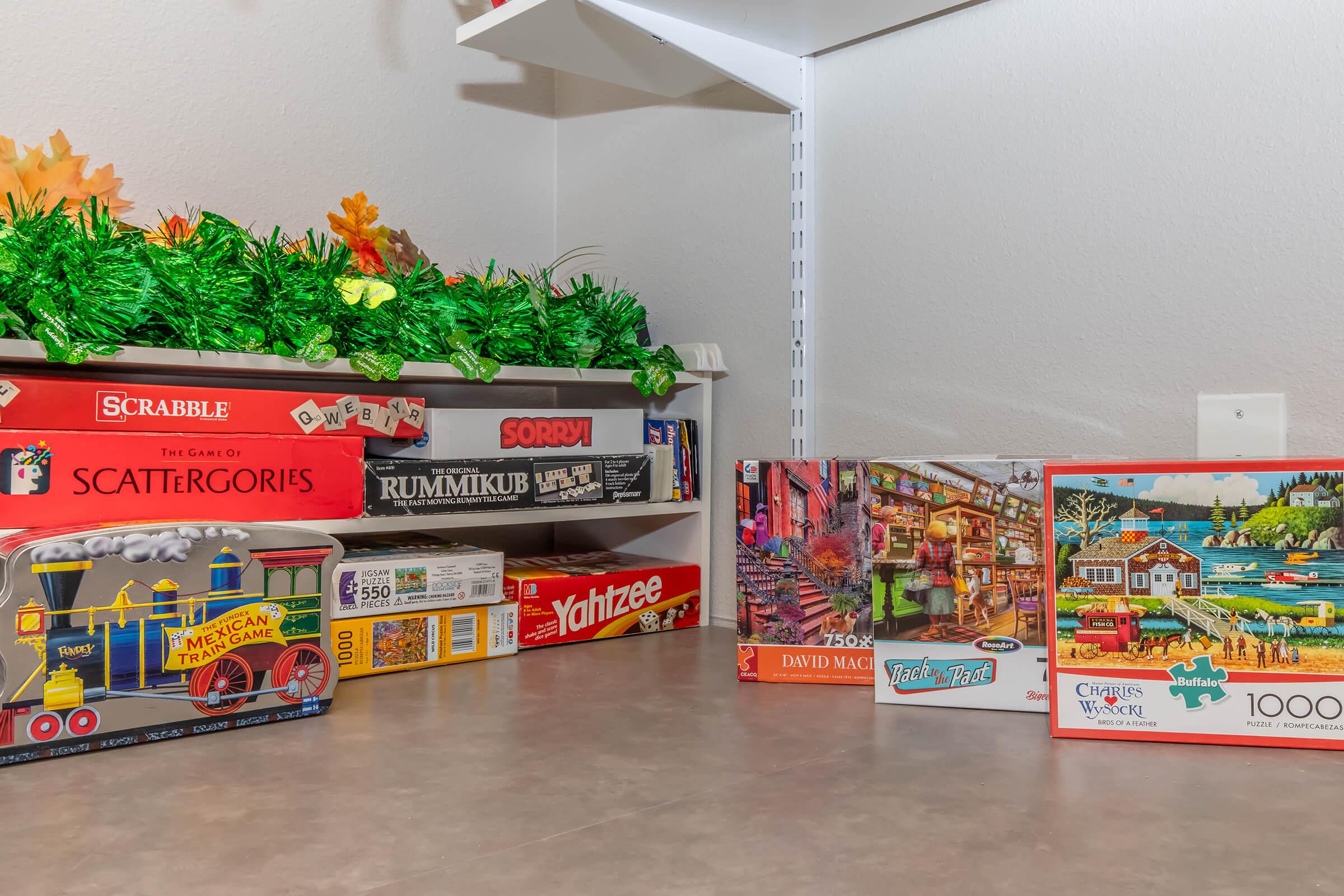 A shelf displaying various board games and puzzles, including Scrabble, Sorry, Rummikub, and Yazzee. There are also colorful puzzle boxes, featuring scenic illustrations and themes. The shelf is decorated with green foliage and autumn leaves, creating a cozy atmosphere.