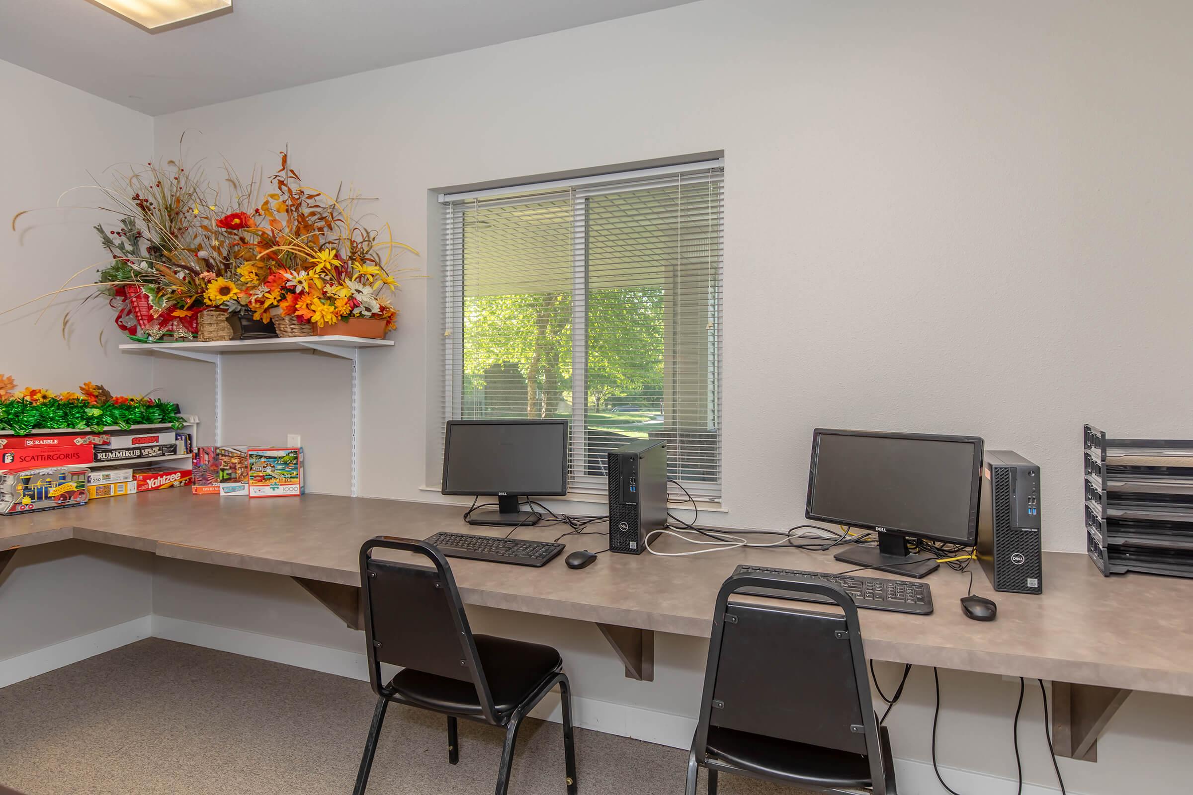 A workspace featuring two desktop computers with monitors and keyboards on a gray table. There are black chairs for seating. A window with blinds offers natural light, and a shelf above the table holds colorful faux flowers and board games. The walls are light-colored, creating a bright and tidy environment.