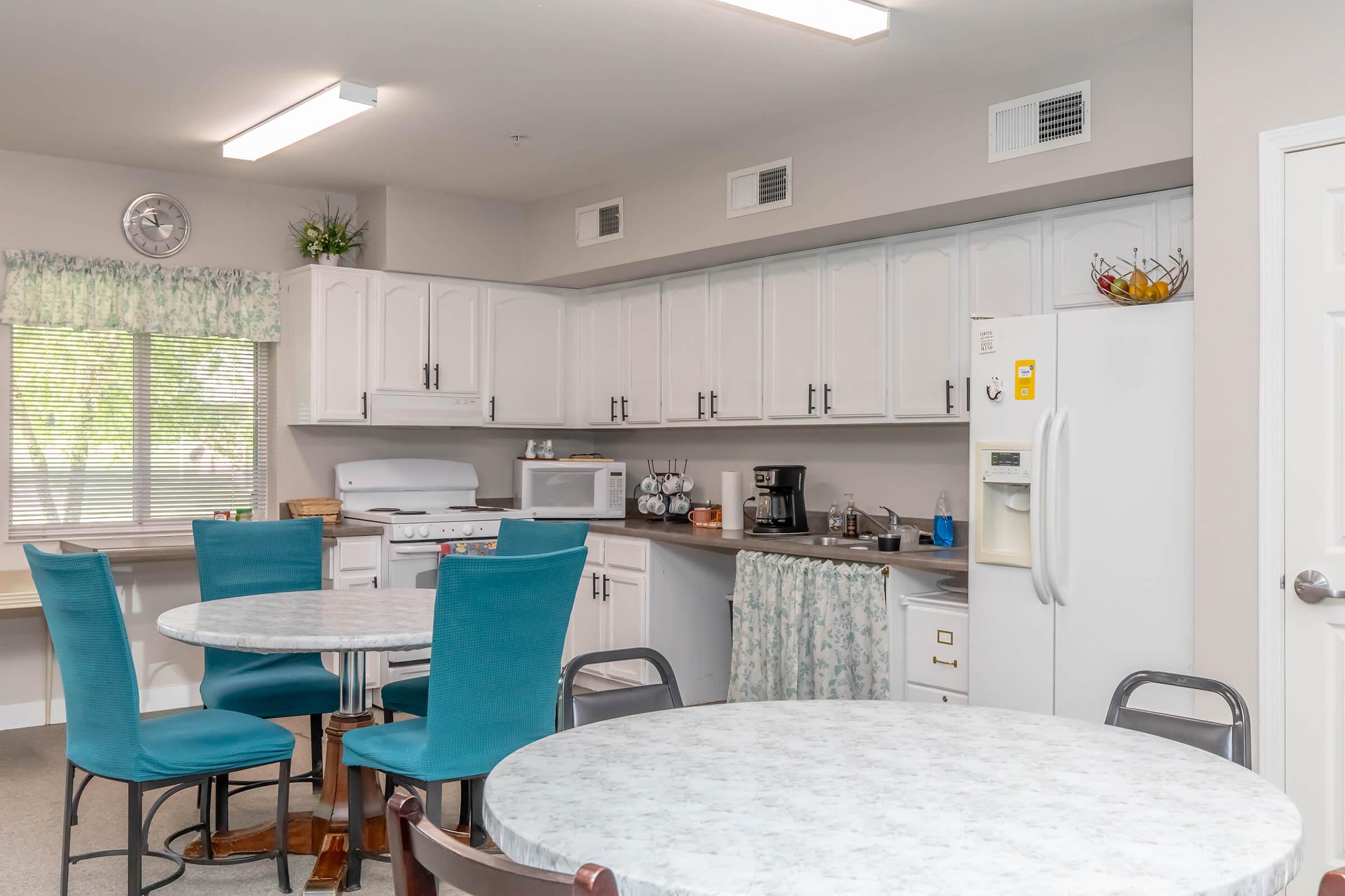 A well-lit kitchen and dining area with two round tables and teal chairs. The kitchen features white cabinets, a stove, microwave, and refrigerator. Natural light enters through a window, enhancing the inviting atmosphere. Decor includes a small fruit basket and colorful curtains.