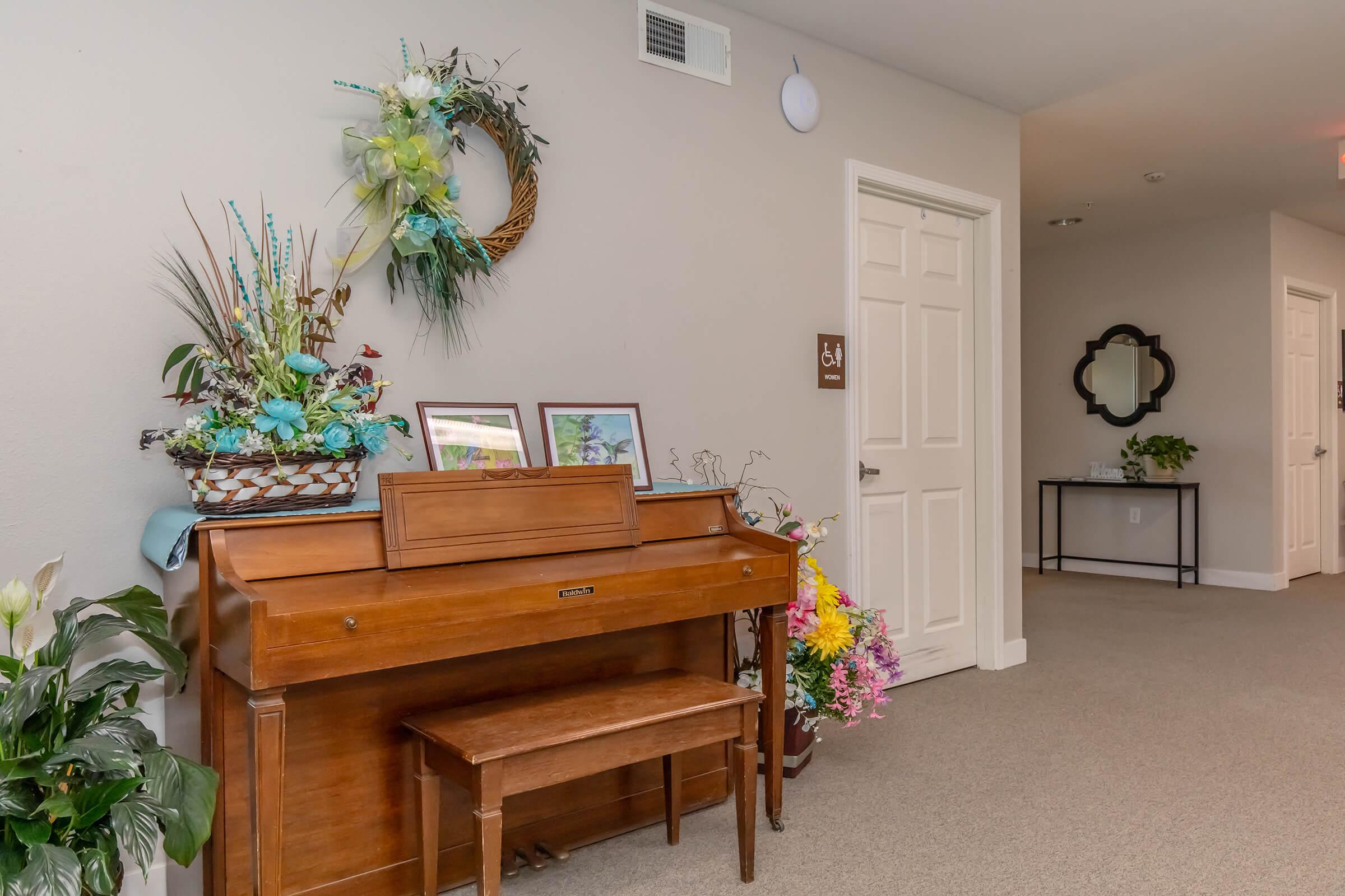 A wooden piano with a matching bench sits in a hallway decorated with plants and floral arrangements. There are framed pictures on the piano, and a wreath hangs on the wall nearby. A door leads to another room, and a mirror is visible on a side table in the background.