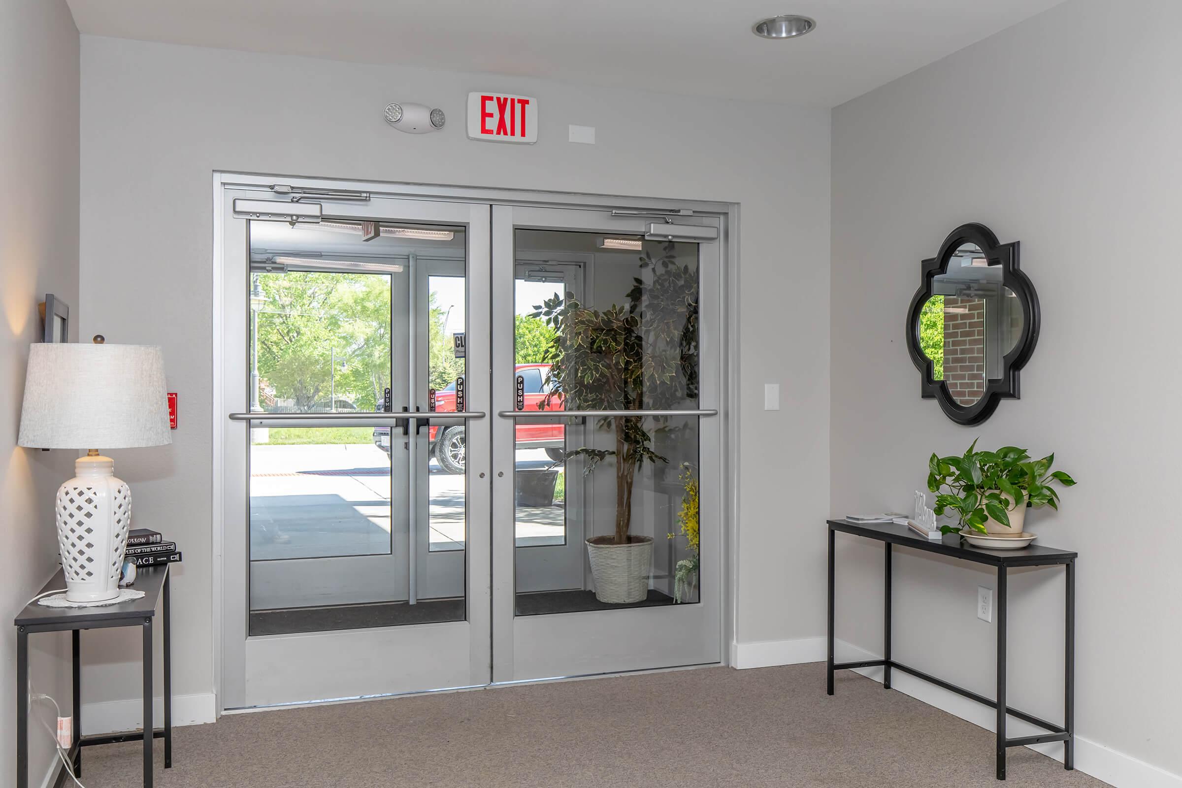 A modern entrance area with double glass doors leading outside. On the left, a lamp sits on a small table, and on the right, a potted plant decorates another table. A round mirror is mounted on the wall, and the space is decorated in neutral colors with natural light coming through the doors. An exit sign is visible above the doors.
