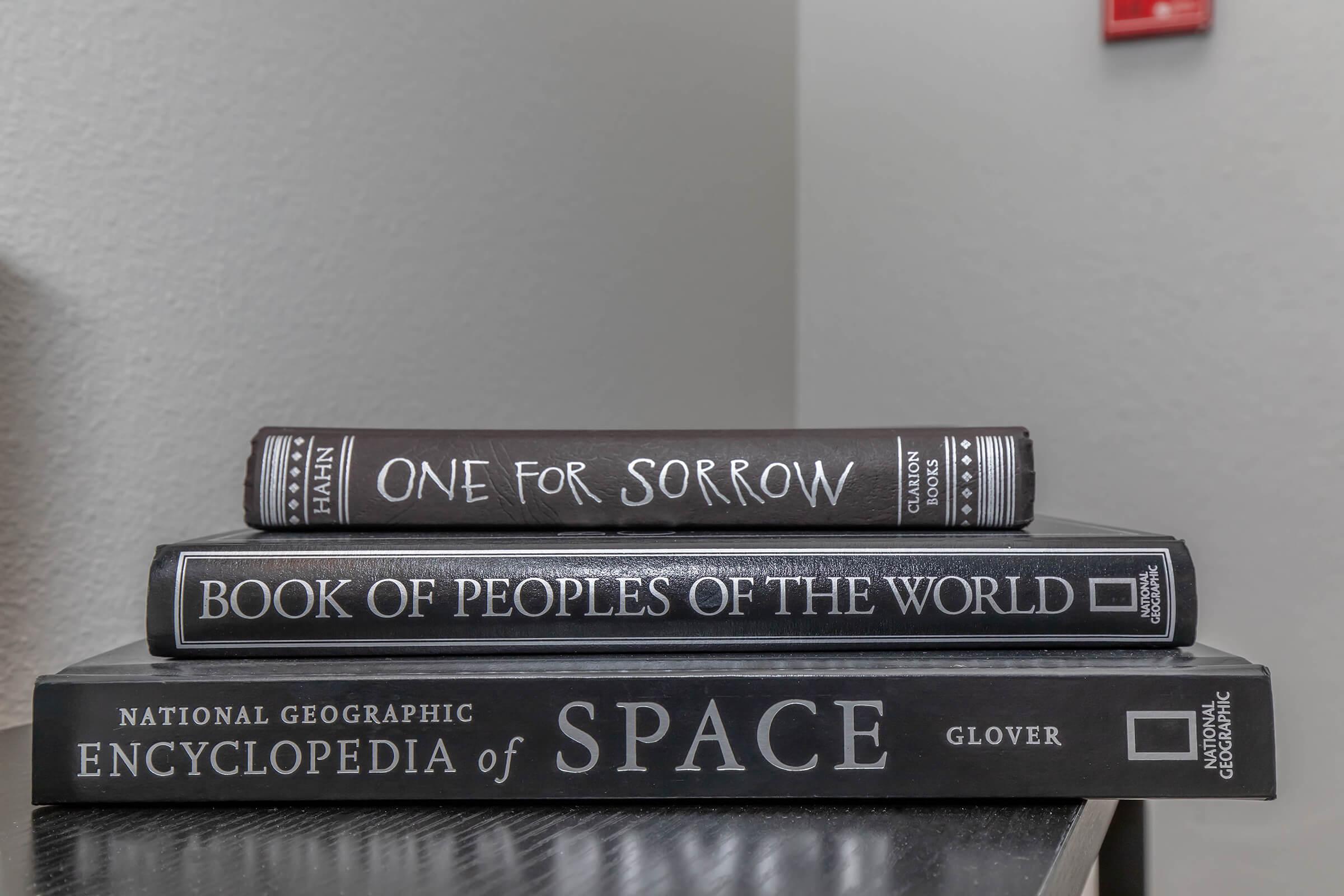 Three stacked books on a gray surface. The top book is titled "ONE FOR SORROW," the middle book is "BOOK OF PEOPLES OF THE WORLD," and the bottom book is "NATIONAL GEOGRAPHIC ENCYCLOPEDIA of SPACE" by Glover. The background is a plain wall, and there is a red object partially visible on the wall.