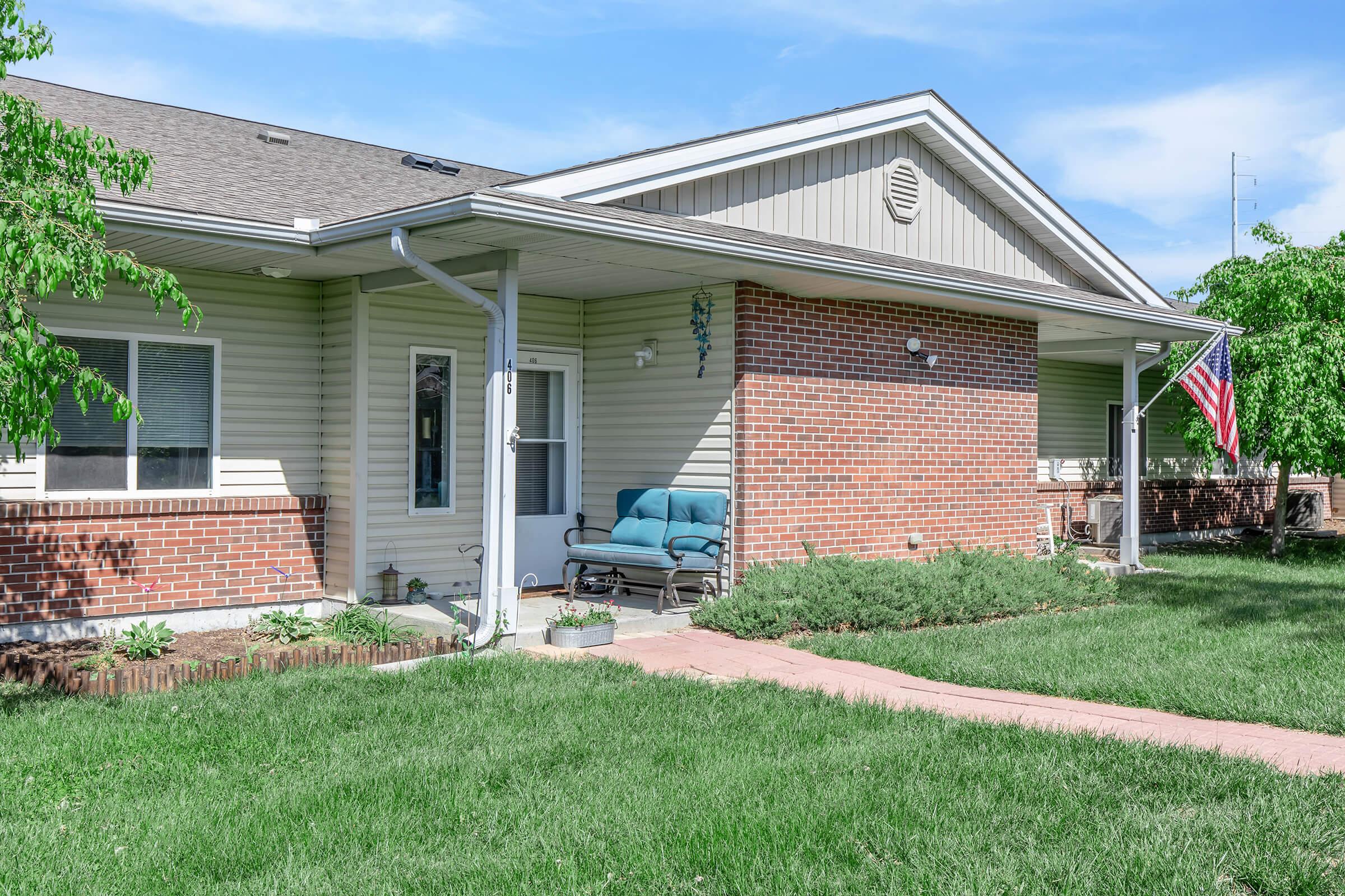 A front view of a single-story house featuring a brick facade and light-colored siding. There is a porch with blue seating, surrounded by green grass and small plants. An American flag is displayed on one side of the house. The sky is clear with a few clouds.