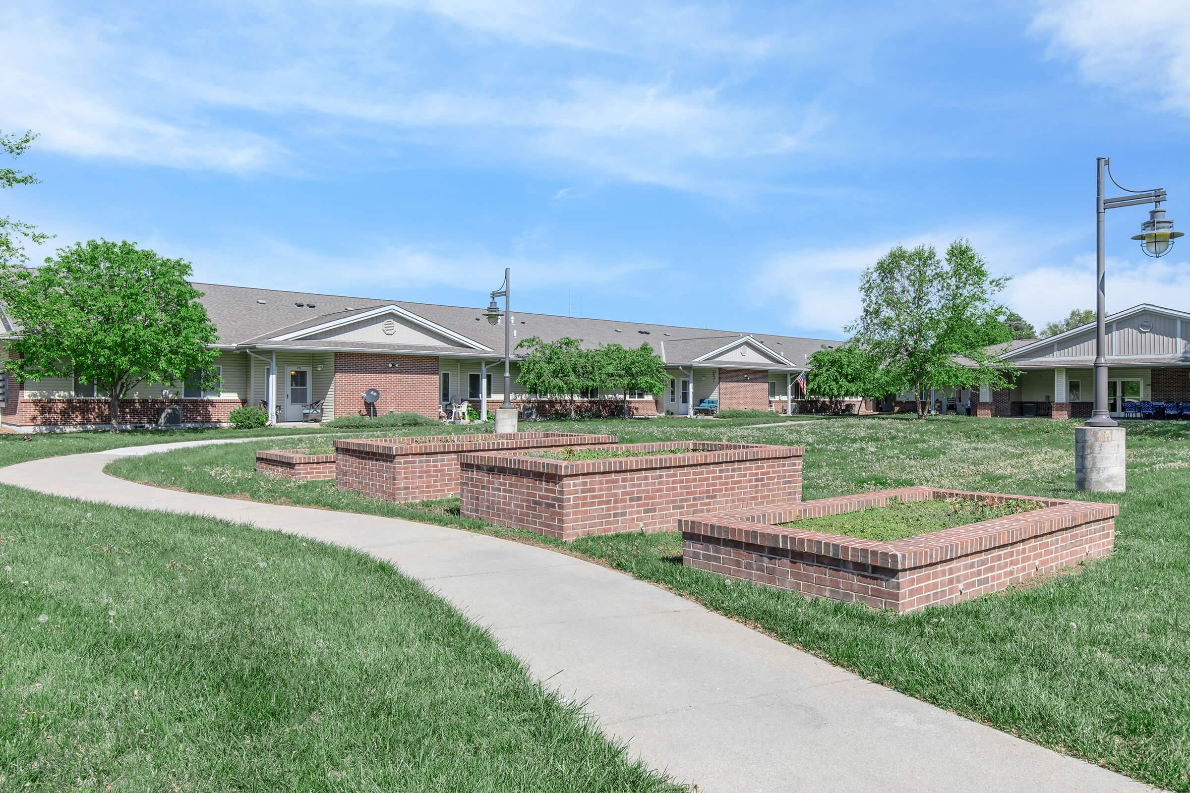 A well-maintained outdoor area featuring a paved walkway winding through green grass. Alongside the path are raised planting beds made of brick, with several trees and shrubs in the background, leading to a series of residential buildings under a clear blue sky.