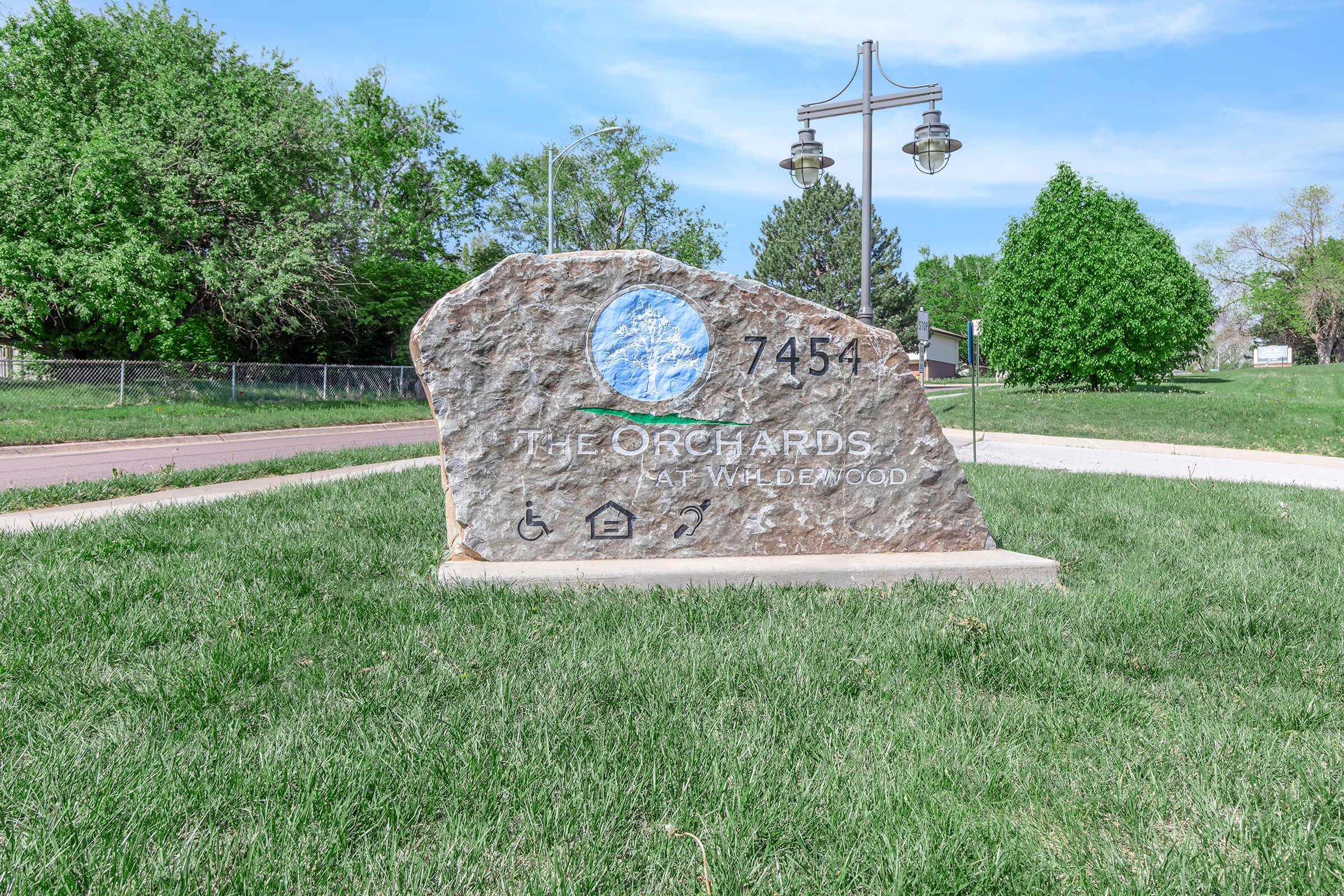 A large stone monument displays the name "J.E. Orchards" along with the address "745-". The sign features a graphic of a blue circle and symbols indicating accessibility. The backdrop includes a grassy area, trees, and a lamppost under a clear blue sky.