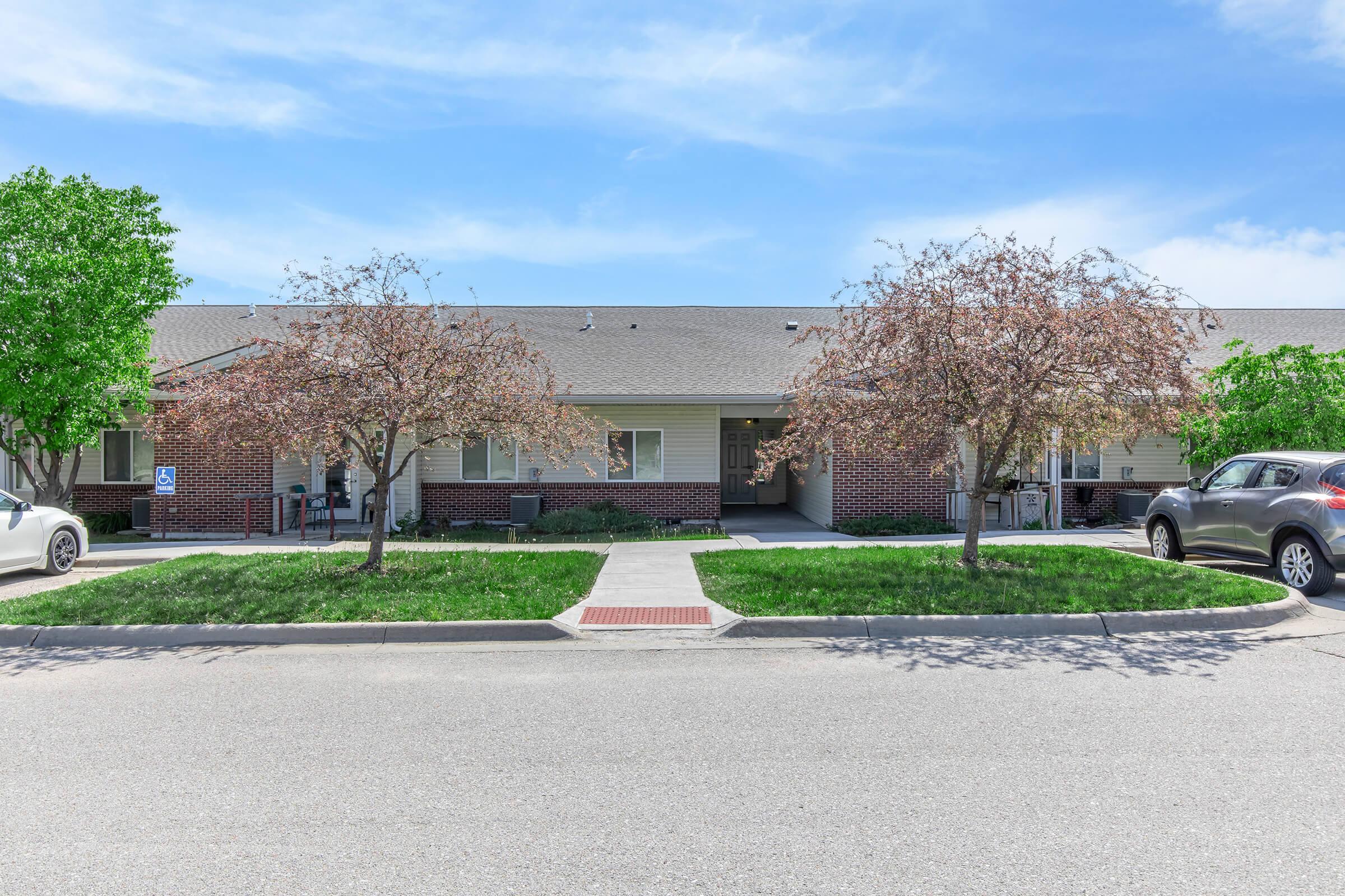 A single-story residential building with a landscaped front yard featuring two small trees. There are parked cars along the street and a concrete pathway leading to the entrance. The building has several windows and is surrounded by green grass under a blue sky.