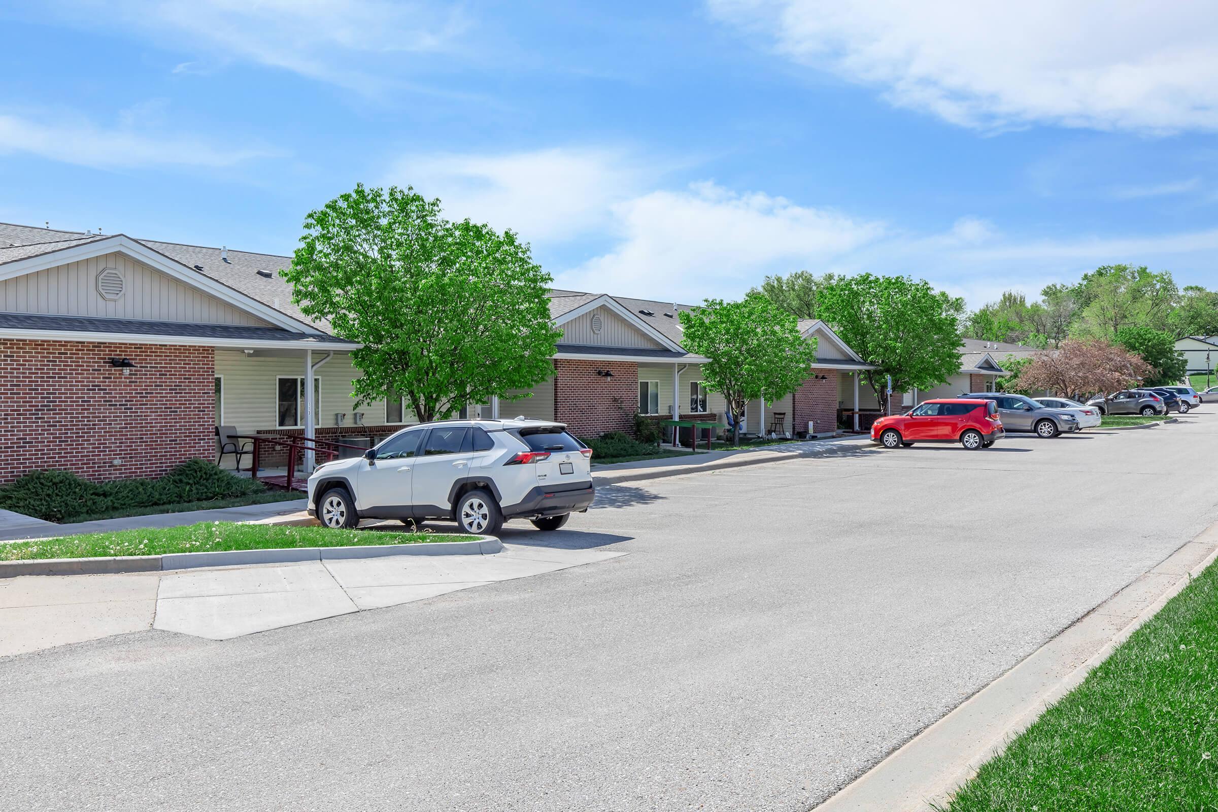 A quiet residential street featuring several one-story brick houses with green foliage and trees. Parked cars, including a white SUV and a red vehicle, are visible along the curb. The sky is clear with a few clouds, suggesting a pleasant day.