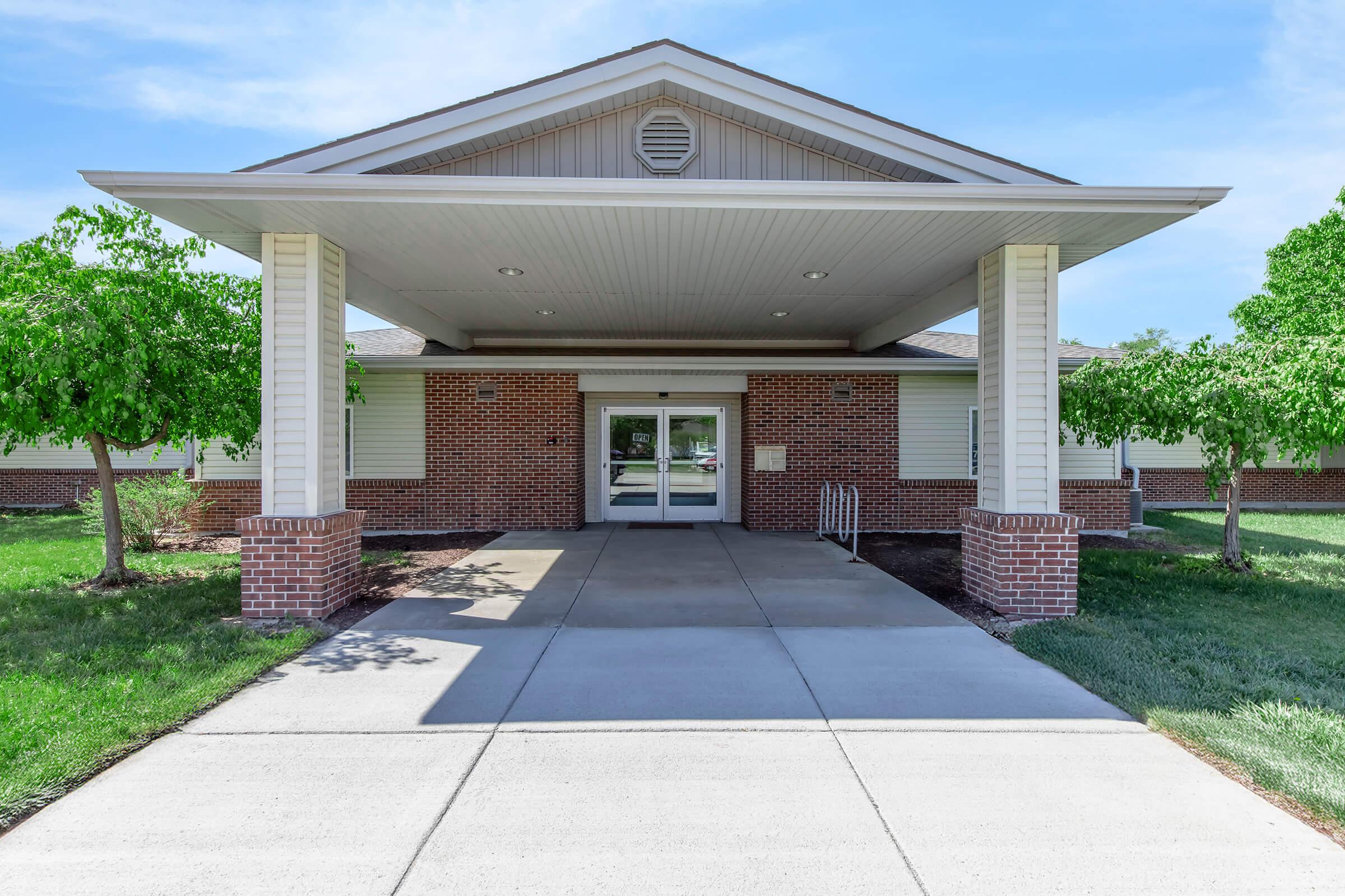 Front view of a modern brick building featuring a large entrance with a covered porch, concrete walkway, and landscaped greenery on either side. The building is set against a blue sky with minimal clouds.