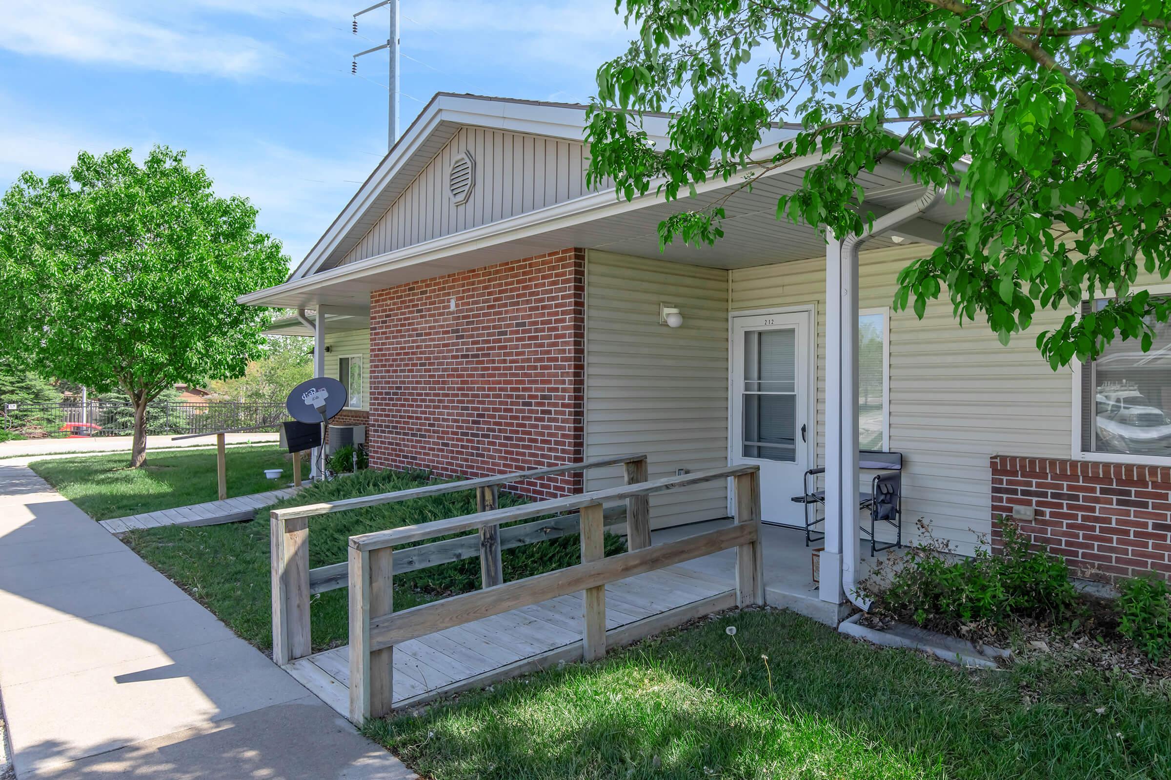 A single-story building with a light-colored exterior and a brick section, featuring a wheelchair-accessible ramp leading to the entrance. The surrounding area has green grass and trees under a clear blue sky. A satellite dish is visible on the side of the building.