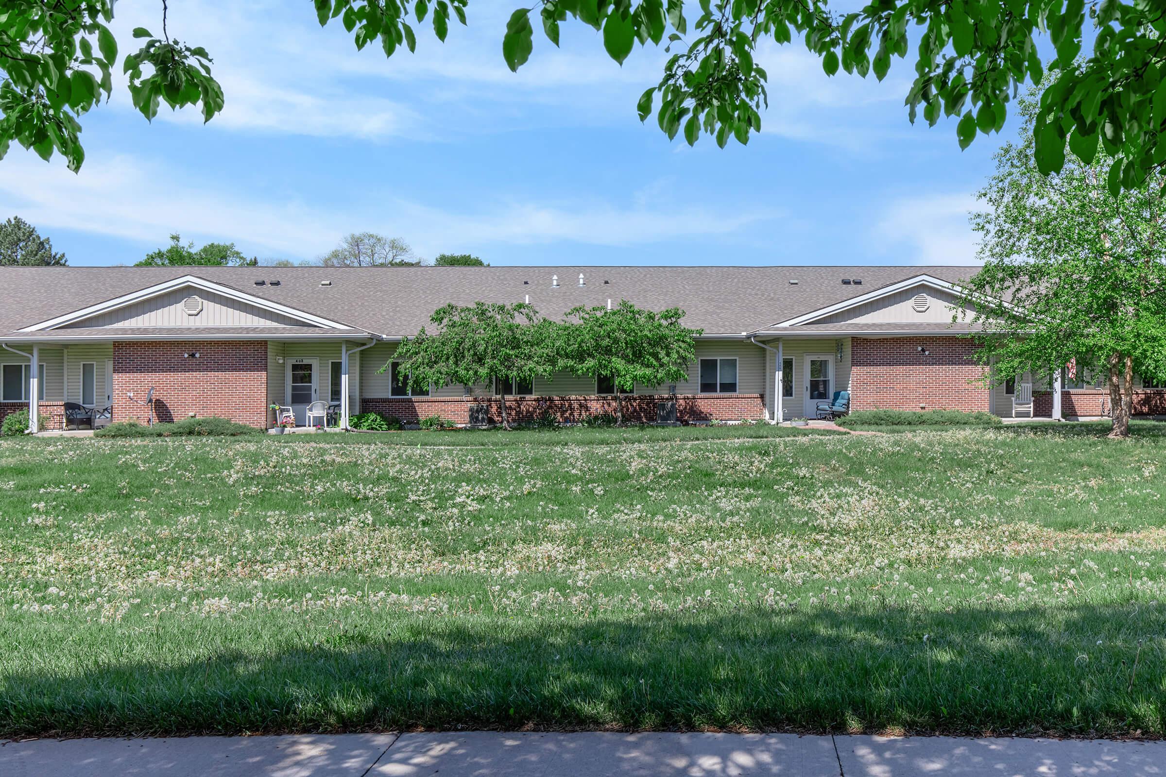 A row of brick residential buildings with white trim, featuring porches and chairs. The foreground shows a grassy area dotted with small white flowers. Trees frame the scene, under a clear blue sky.