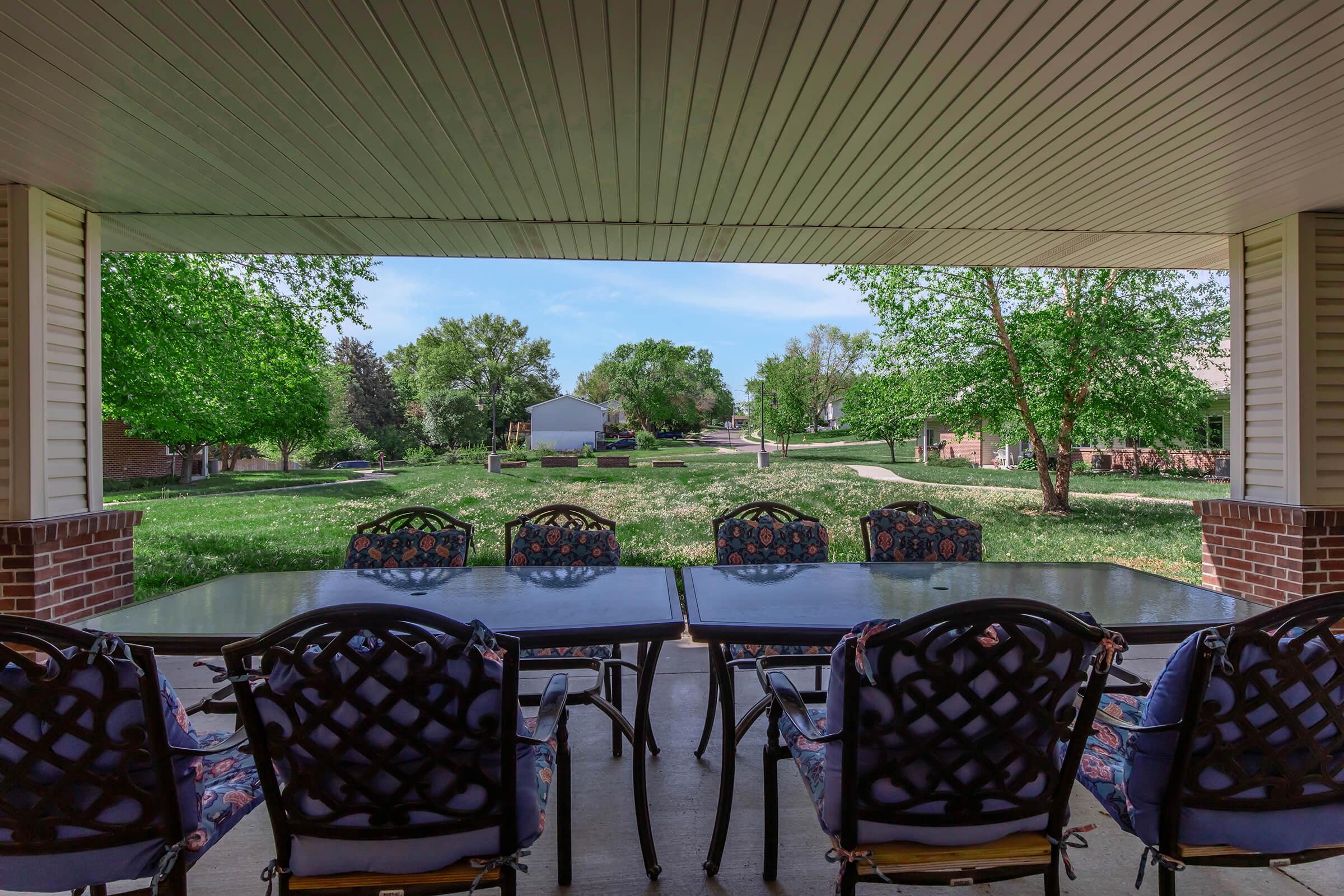 View from a covered patio featuring a table and chairs, overlooking a grassy area with trees and a distant house. The outdoor space is bright and inviting, with clear blue skies and well-maintained landscaping.
