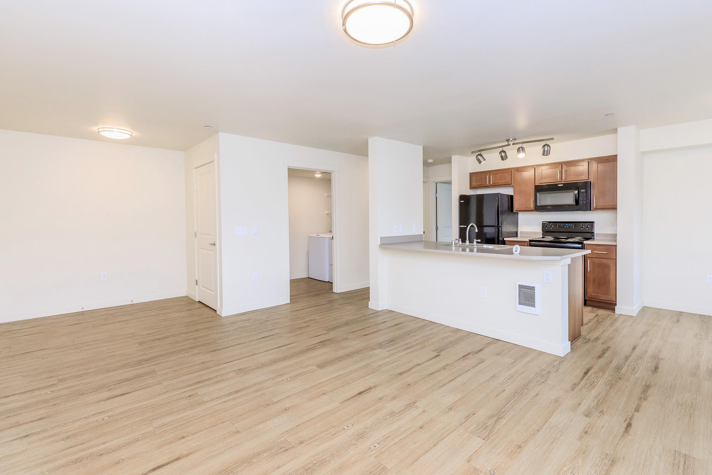 Modern open-concept living area featuring vinyl flooring, neutral-colored walls, and a well-equipped kitchen with dark wood cabinetry and stainless steel appliances. A laundry area is visible through a doorway. Soft lighting fixtures enhance the spaciousness of the room.