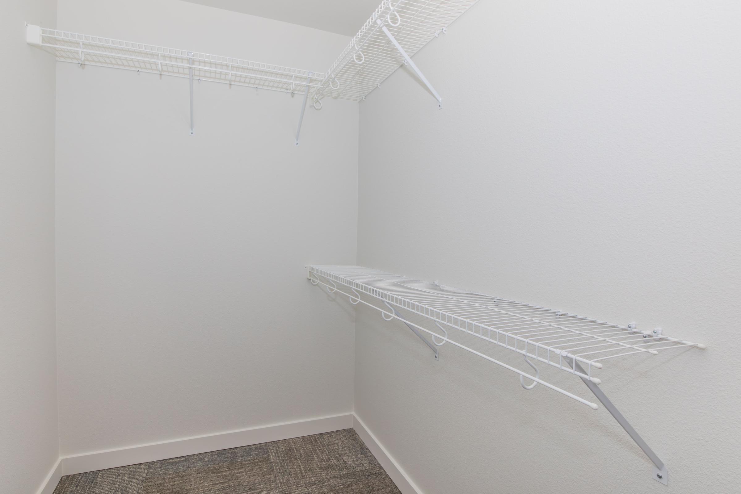 Empty closet space with white wire shelving along the walls, featuring one shelf at the top and another lower shelf. The walls are a light gray color, and the flooring is a neutral carpet. The overall space appears clean and organized, suitable for storage or hanging clothes.