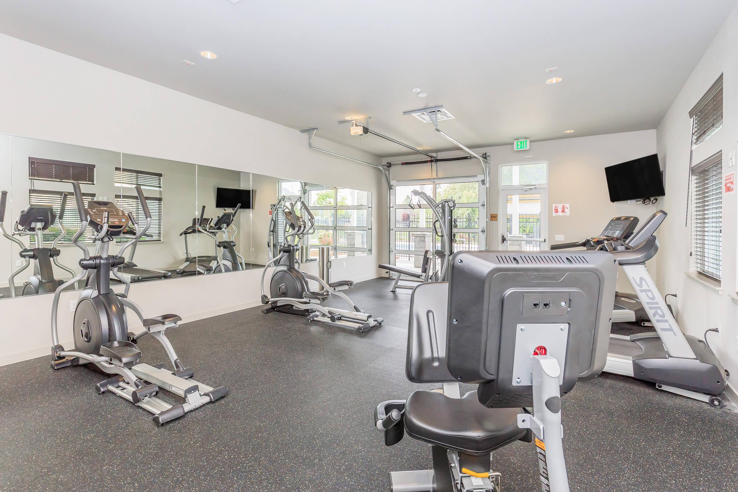Bright, modern gym space featuring various exercise equipment including ellipticals and stationary bikes. Large mirrors line one wall, and there are windows providing natural light. The floor is covered with black rubber material, and there’s a television mounted on the wall.