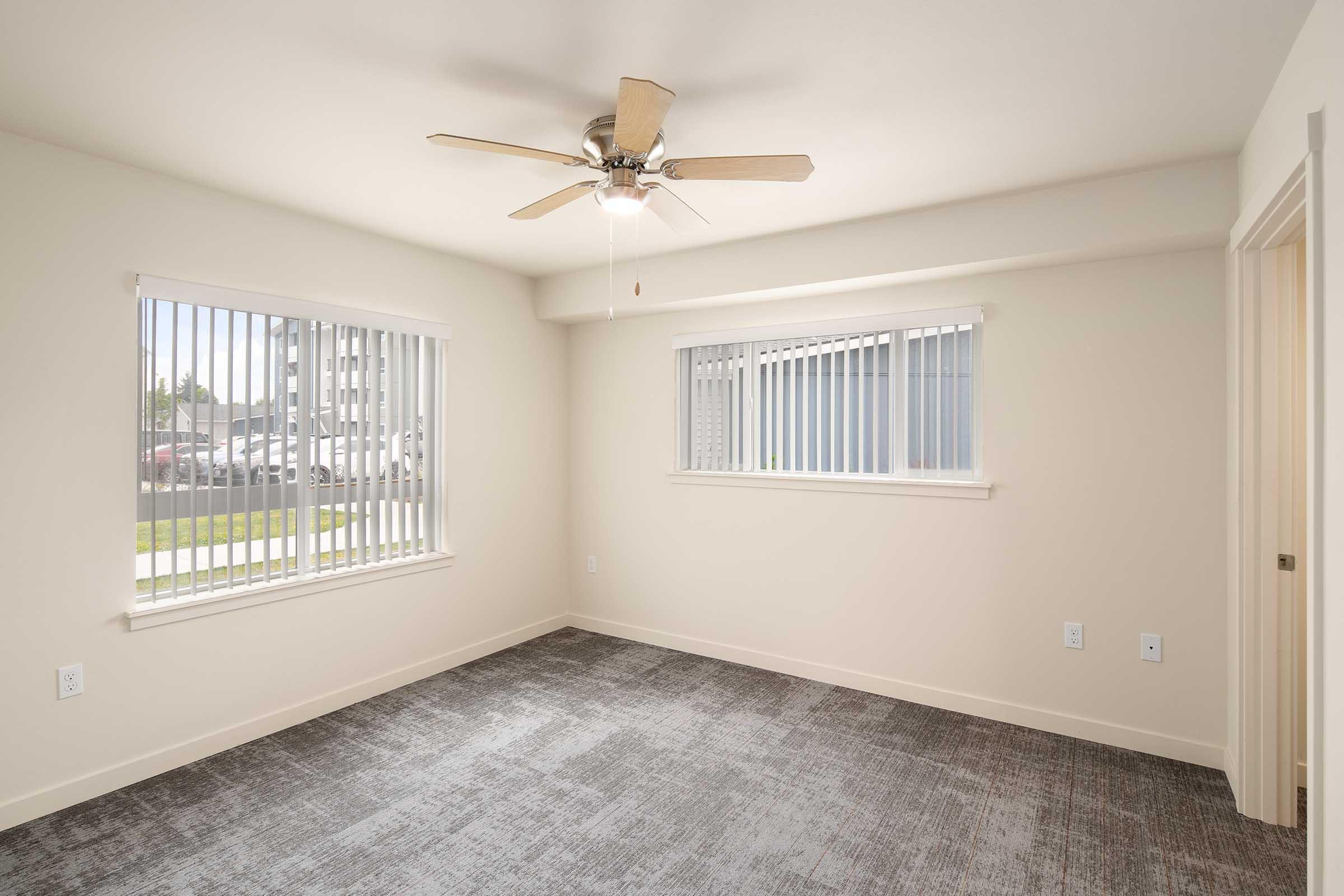 A bright, empty room with light-colored walls and a ceiling fan. Two windows with horizontal blinds allow natural light to enter. The floor is covered with gray carpet, and there are no furnishings. A doorway is visible on the right side of the image, leading to another area.