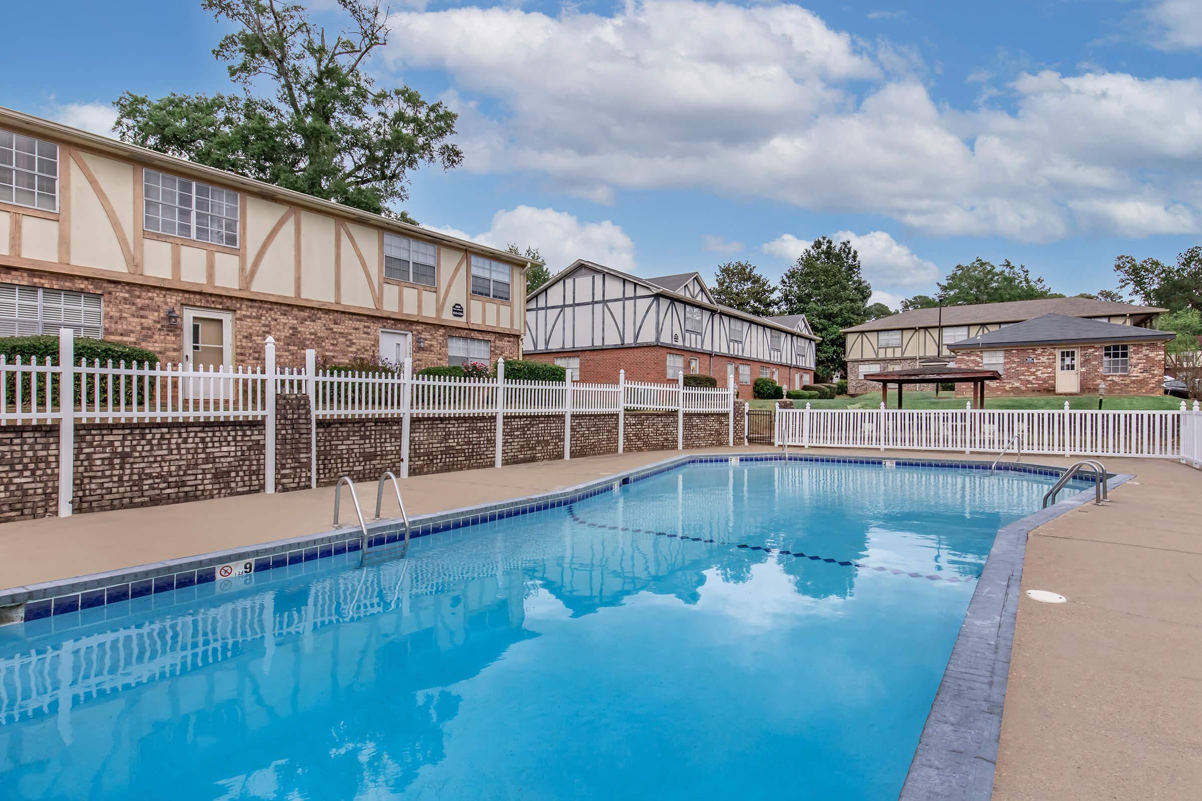 Swimming pool with clear blue water, surrounded by a concrete deck. White picket fencing lines the area, with brick buildings featuring a Tudor architectural style in the background. Lush greenery and trees are present, along with a cloudy sky above.