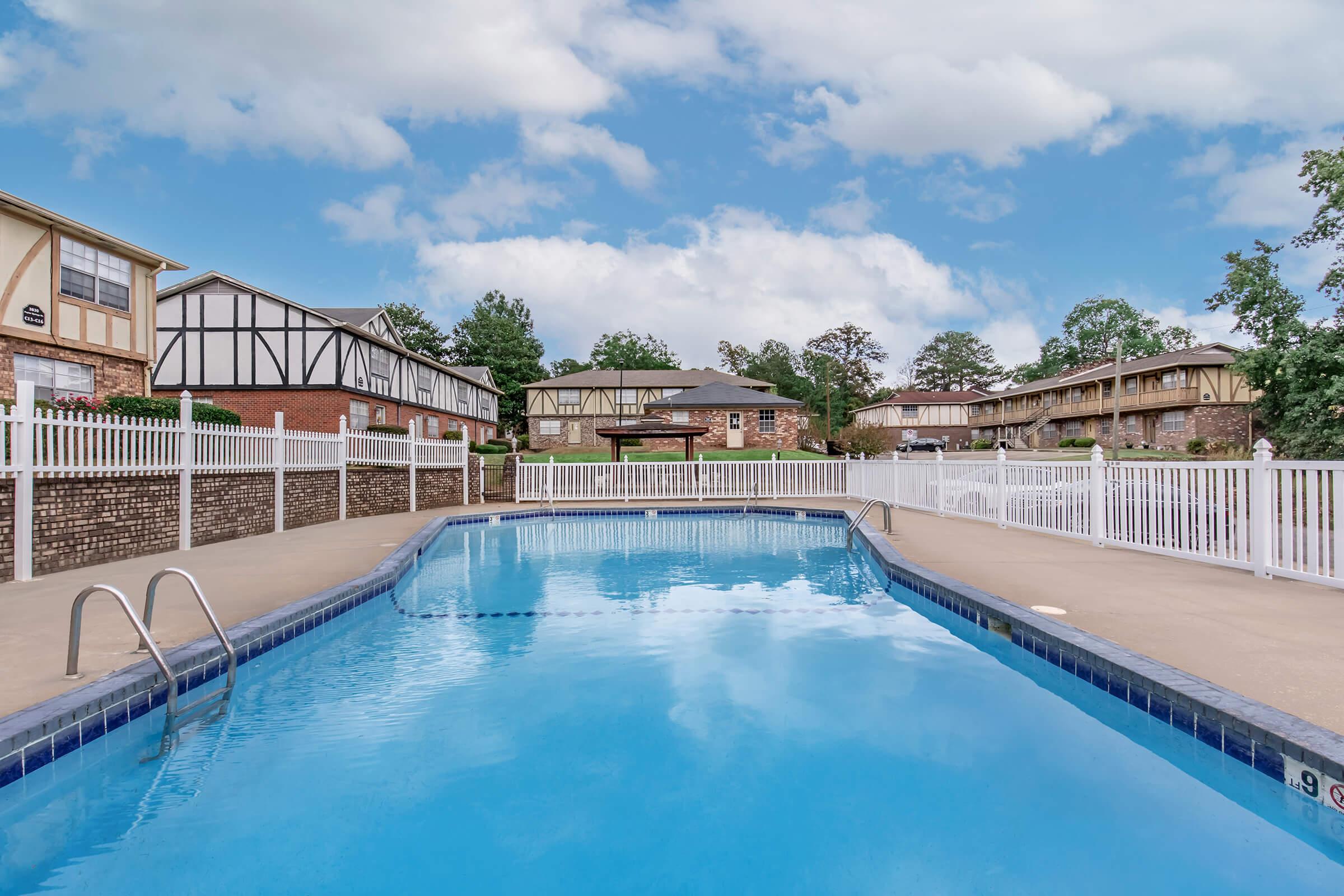 Swimming pool with clear blue water surrounded by a paved deck and white fencing. In the background, there are well-maintained brick apartment buildings and lush greenery under a partly cloudy sky. The area is spacious and inviting, ideal for relaxation and leisure.