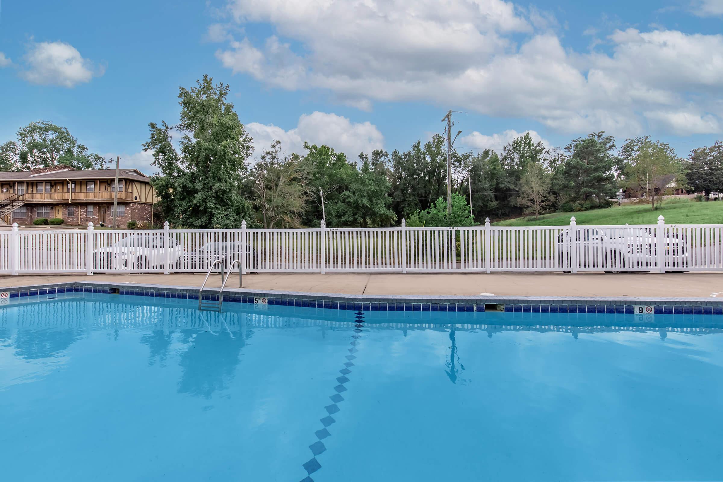 A clear blue swimming pool with white lounge chairs surrounding it. In the background, there's a green lawn and trees, and a wooden building partially visible. The sky is partly cloudy, creating a serene atmosphere.