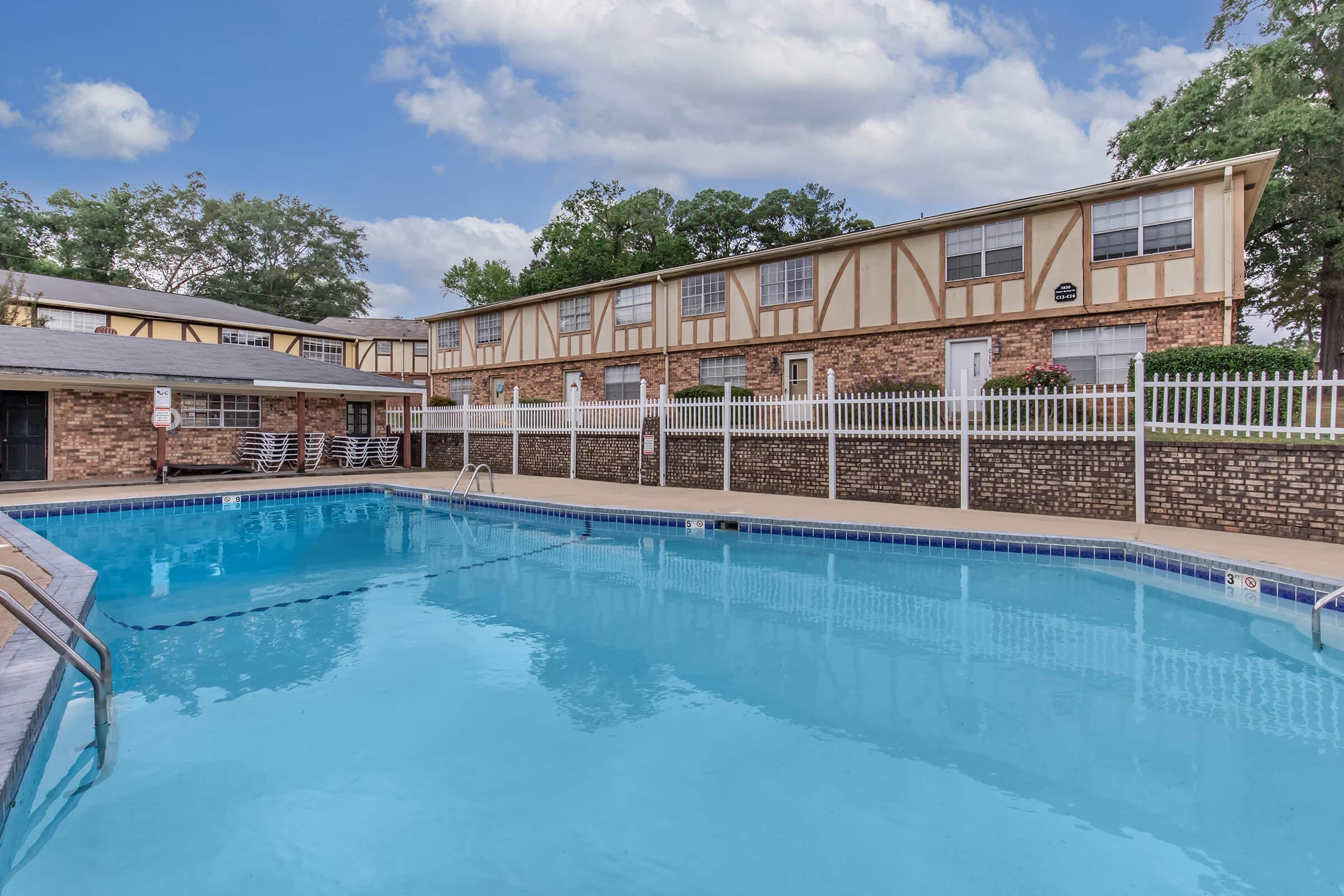 A clear swimming pool surrounded by a fenced area, with a brick building and several trees visible in the background. The pool area features sun loungers and a blue tile border, under a partly cloudy sky. The setting appears inviting and well-maintained.
