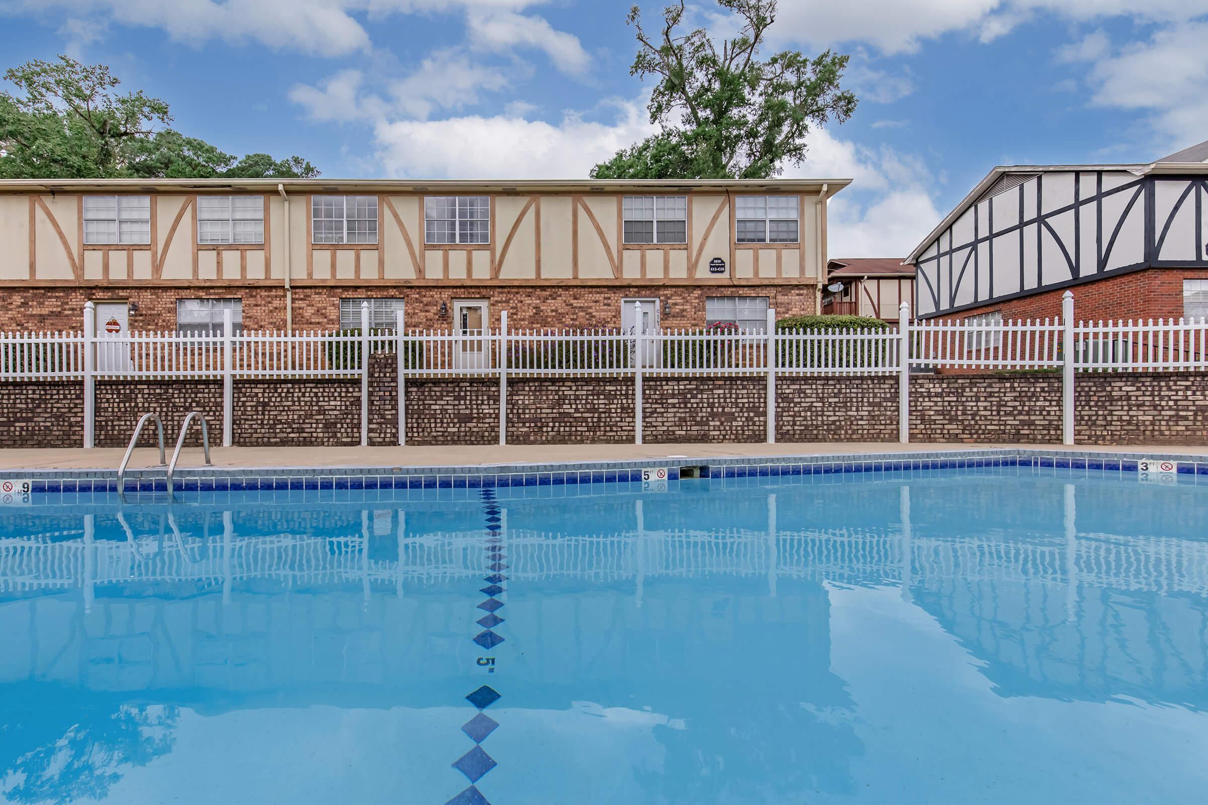 A swimming pool with clear blue water, lined with a white picket fence. In the background, there are residential buildings with brick and wood siding. The sky is partly cloudy, and some trees can be seen in the background. The pool area is well-maintained and inviting.