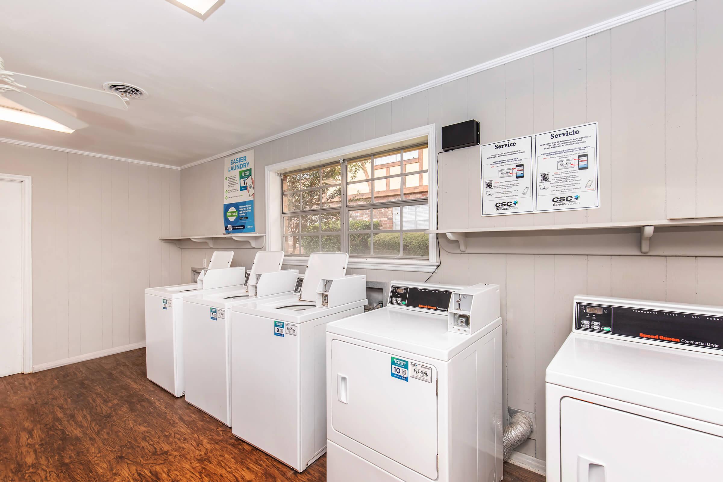 A clean laundry room featuring several stacked white washing machines and dryers, with a large window allowing natural light. The walls are light-colored wood paneling, and there are shelf spaces above the machines. Informational signs about laundry procedures are posted on the walls.