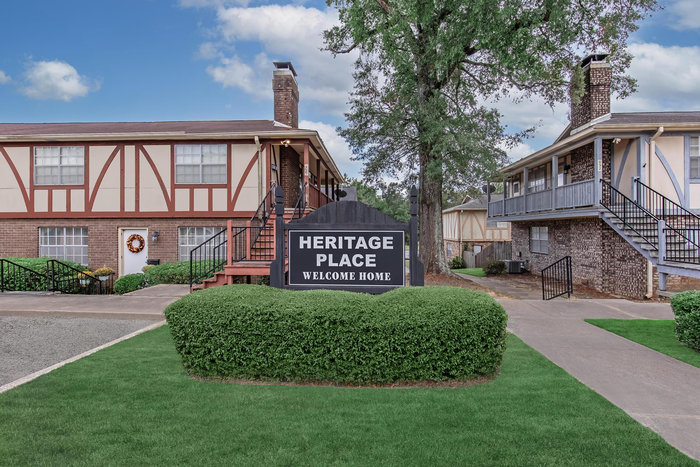 Sign for "Heritage Place" with the text "Welcome Home" in a landscaped area. Two residential buildings are visible, featuring brick and siding exteriors, with staircases leading to entrances. Green grass and trees enhance the inviting atmosphere.