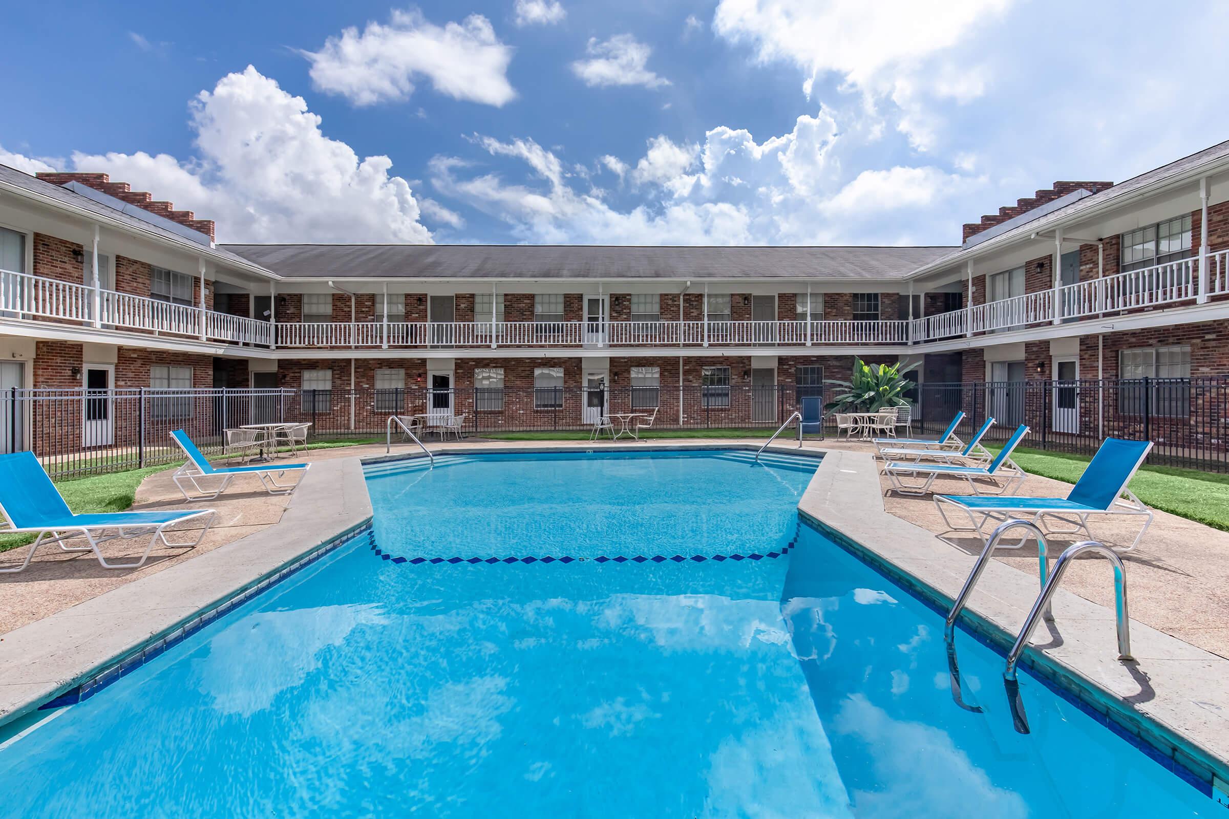A sunny view of a rectangular swimming pool surrounded by lounge chairs, with a brick building in the background featuring multiple balconies. The sky is partly cloudy, and the scene conveys a relaxed atmosphere typical of a motel or apartment complex.