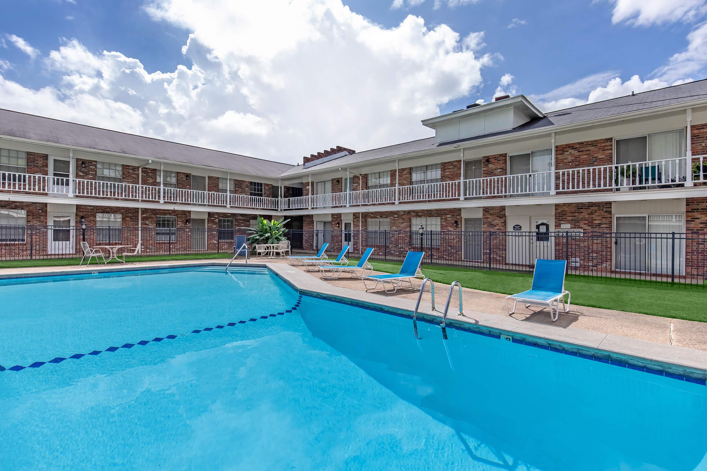 A clear, inviting swimming pool surrounded by lounge chairs, situated in the courtyard of a two-story brick building. The sky is partly cloudy, enhancing the bright and relaxing atmosphere of the area. Grass borders the pool, creating a welcoming environment for guests.