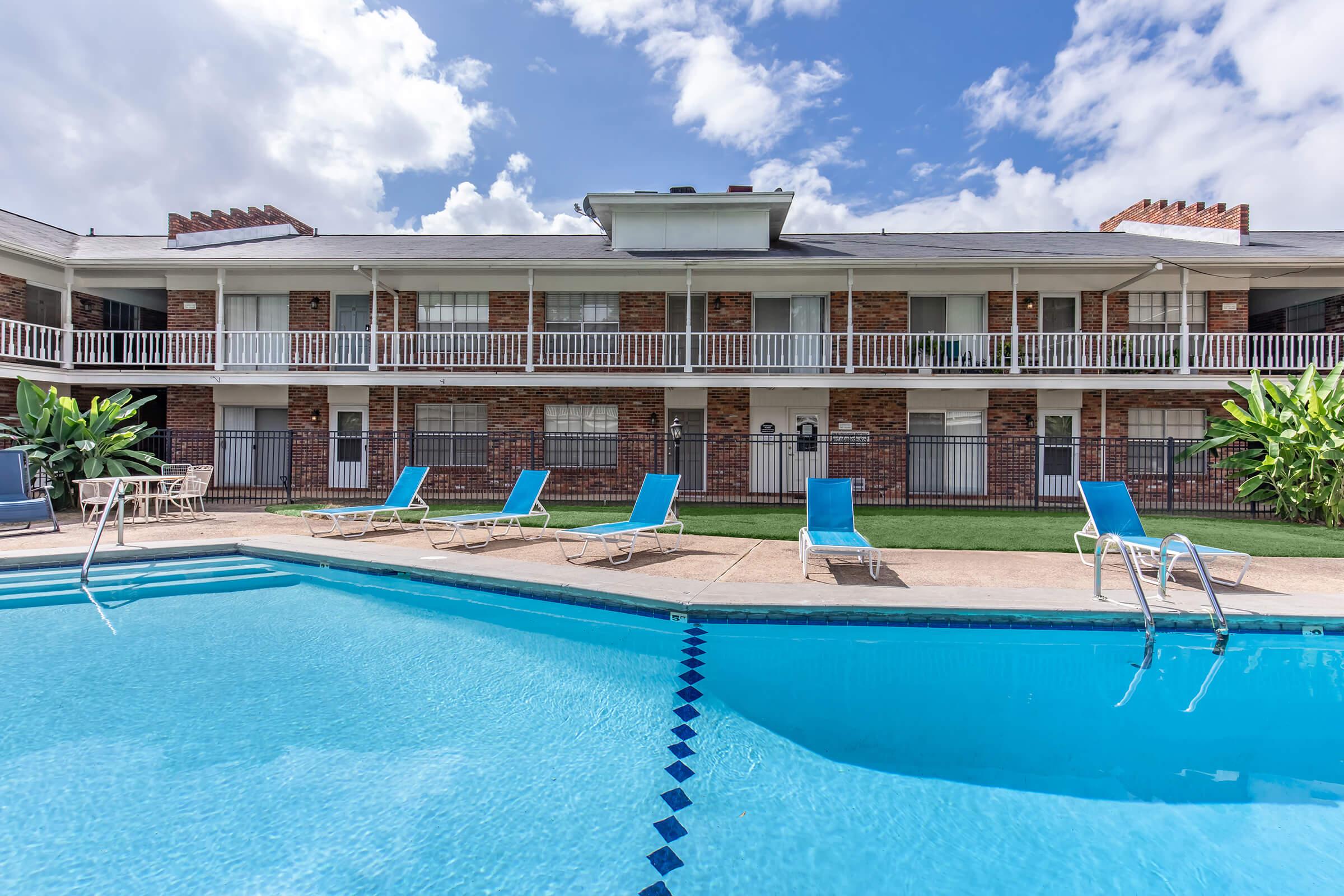 Swimming pool surrounded by lounge chairs, with a brick building featuring multiple balconies in the background. The sky is partly cloudy, creating a bright, inviting atmosphere. The grass around the pool adds a touch of greenery to the scene.