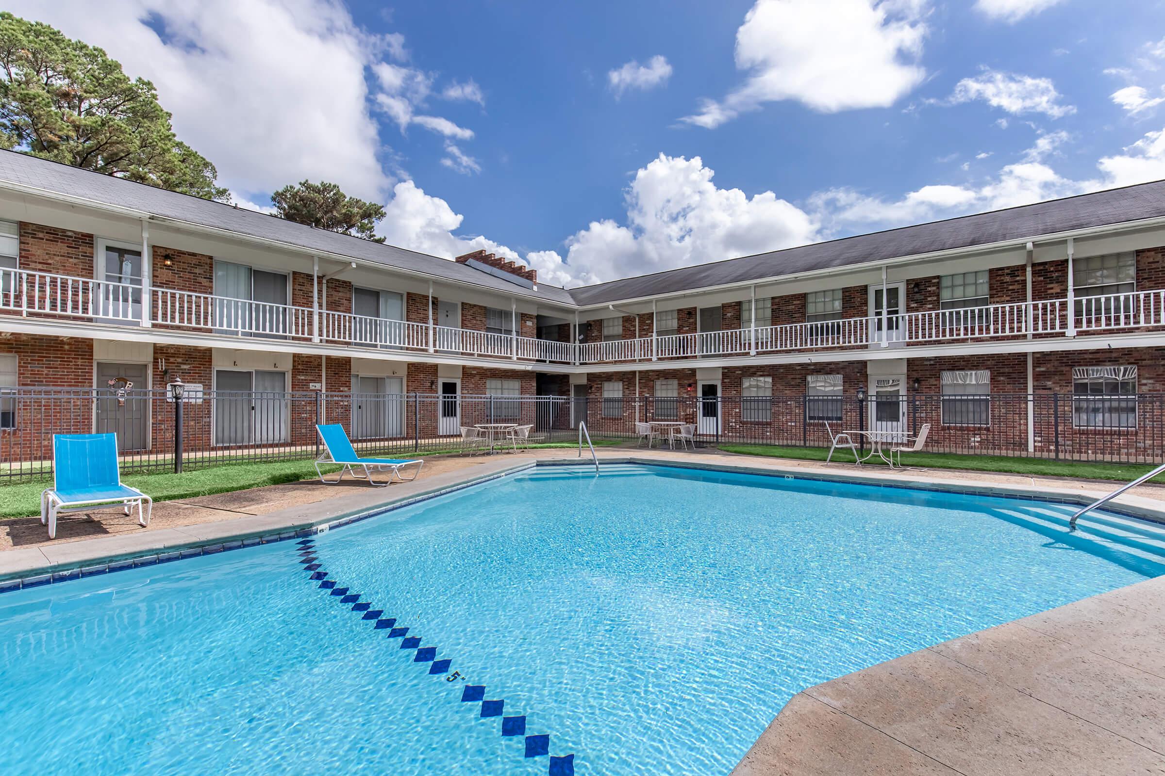 A clear blue swimming pool surrounded by lounge chairs, located in the courtyard of a multi-story brick apartment complex. The building features white railings and balconies, with a bright sky and scattered clouds in the background. The scene conveys a relaxing atmosphere.