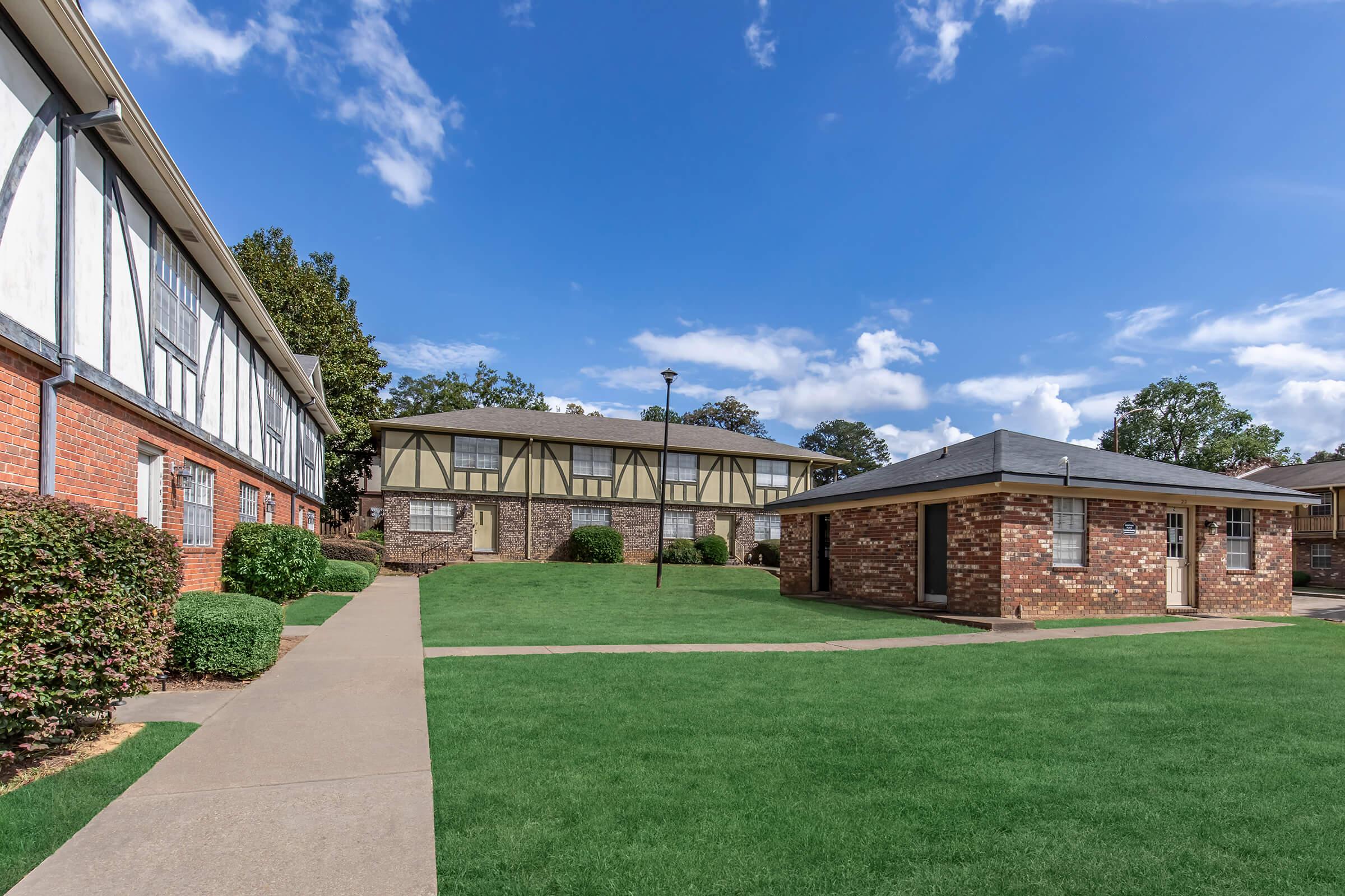 A landscaped courtyard featuring several residential buildings. The foreground has neatly trimmed grass and concrete pathways, while the background displays two distinct building styles: one brick with windows and another with a half-timbered design under a bright blue sky with scattered clouds.