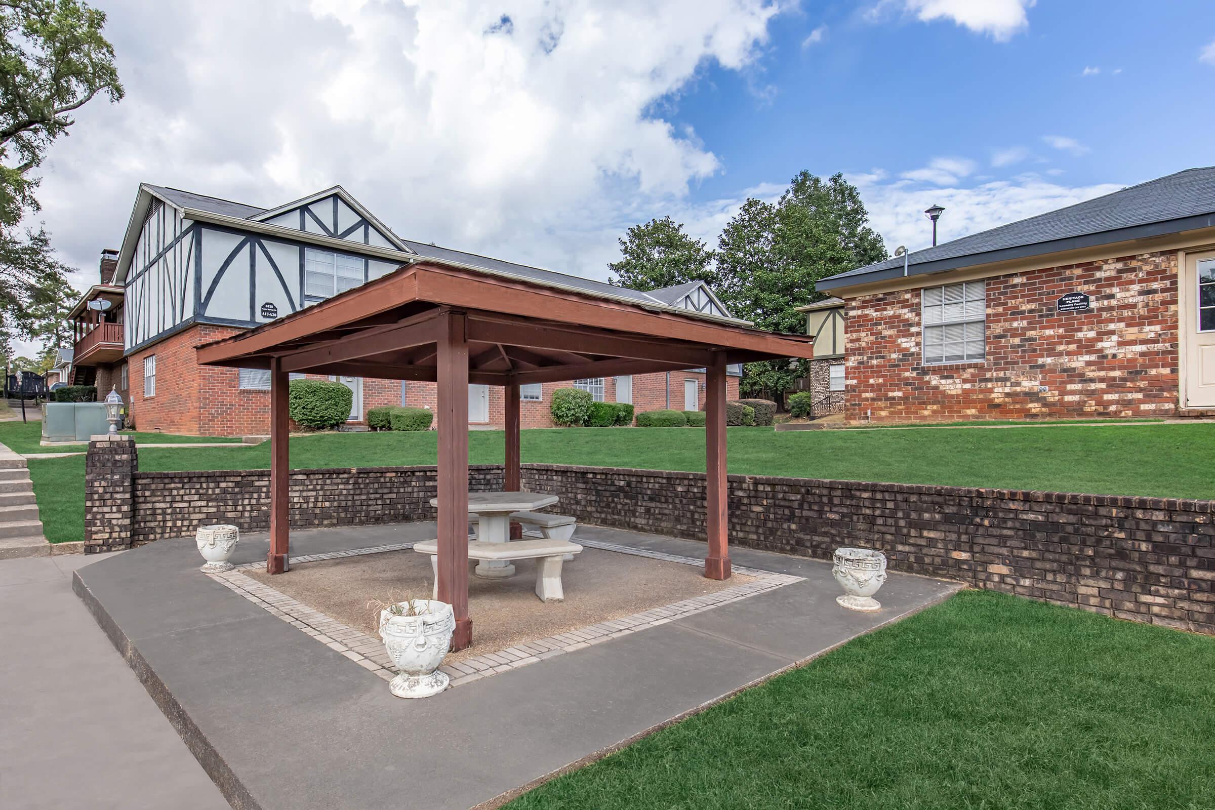 A shaded gazebo with a stone table and benches, surrounded by manicured green lawns and brick buildings. The sky is partly cloudy, and the area is well-maintained, suggesting a pleasant outdoor space for relaxation. Decorative white planters are placed near the gazebo.