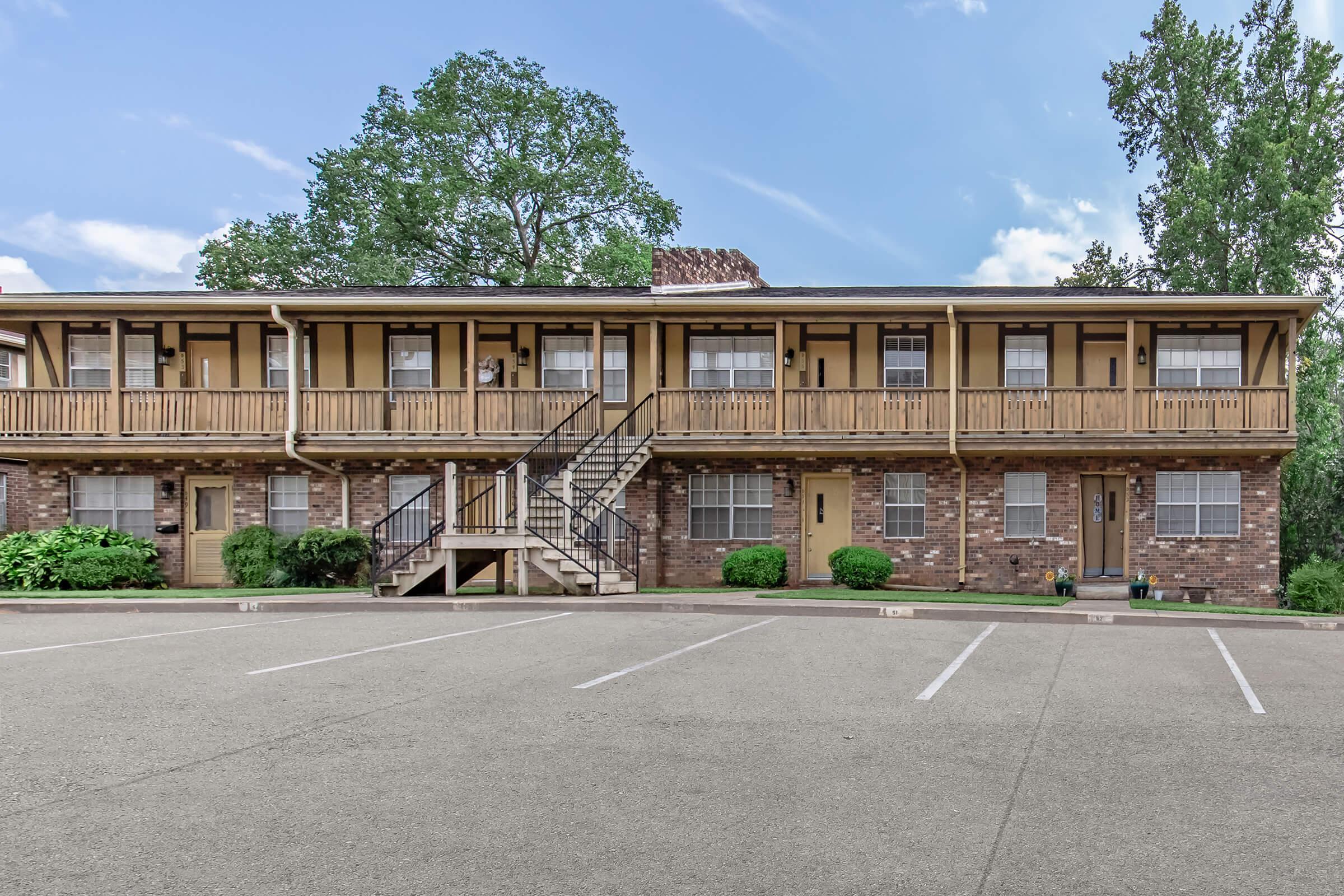 A two-story apartment building with a wooden balcony on each level, surrounded by trees and greenery. The building features brick and yellow siding, with several doors visible on the ground and upper floors. There is a parking lot in front with designated spaces.