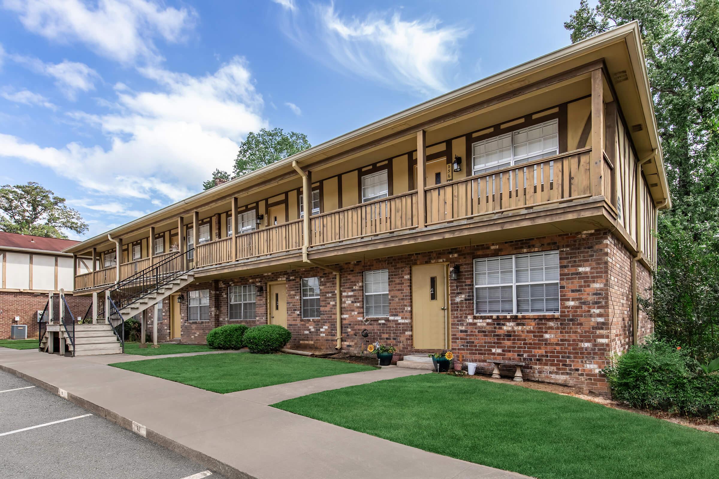 A two-story apartment building with a brick facade and wooden balconies. The building features a staircase leading to the second level, green grass in front, and small shrubs alongside the walkway. The sky is partially cloudy, creating a pleasant atmosphere.